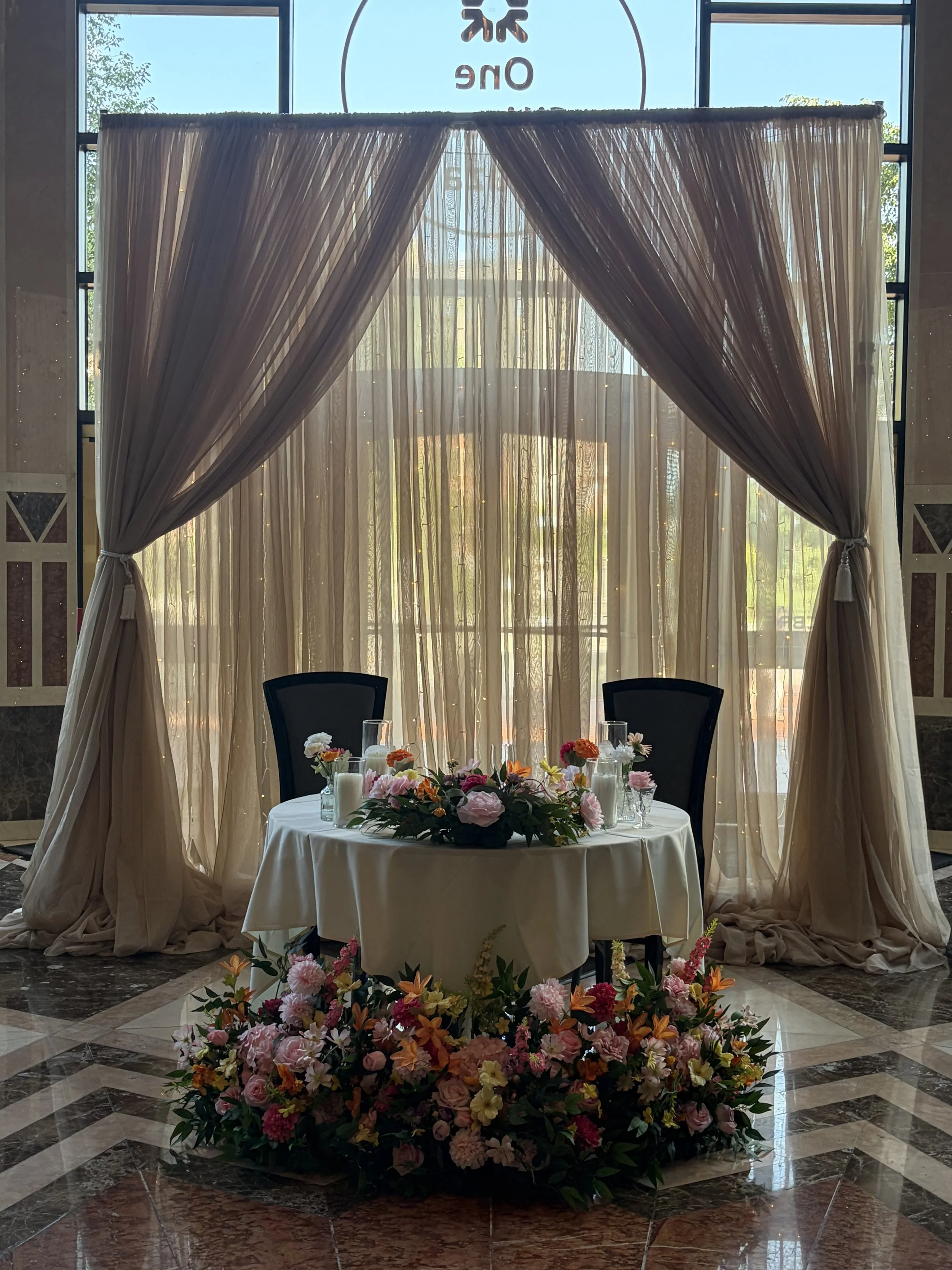 A round table with a white tablecloth and floral arrangements in glass vases, set up for a celebration or event, positioned in front of a large window with beige curtains tied back, and a flower wall backdrop.