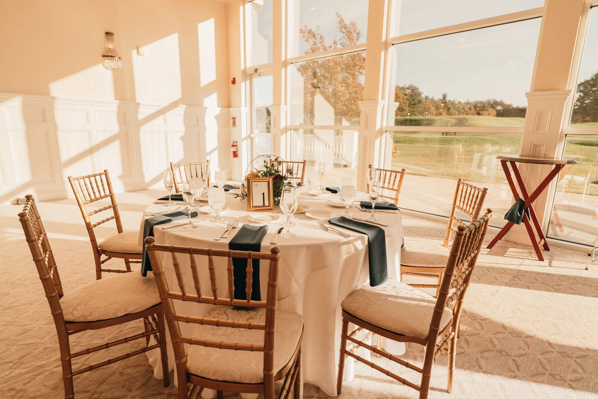 Round banquet table with beige upholstered chairs, set with white tablecloths, black napkins, wine glasses, and a centerpiece, in a sunlit room with large windows and view of a golf course.