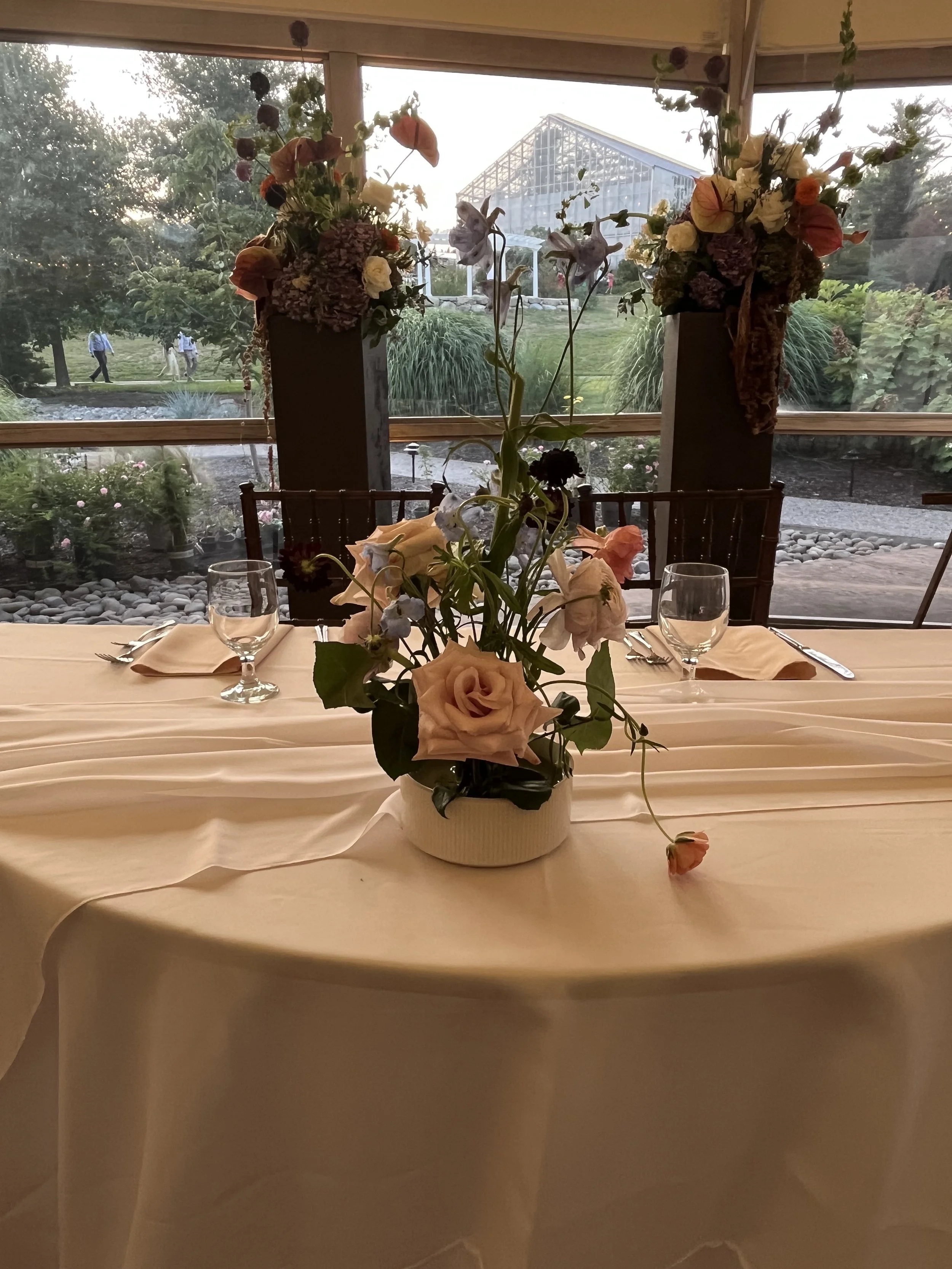 A table with a floral centerpiece featuring roses and other flowers, set with glasses, napkins, and silverware, with a large window showing a garden and greenhouse in the background.