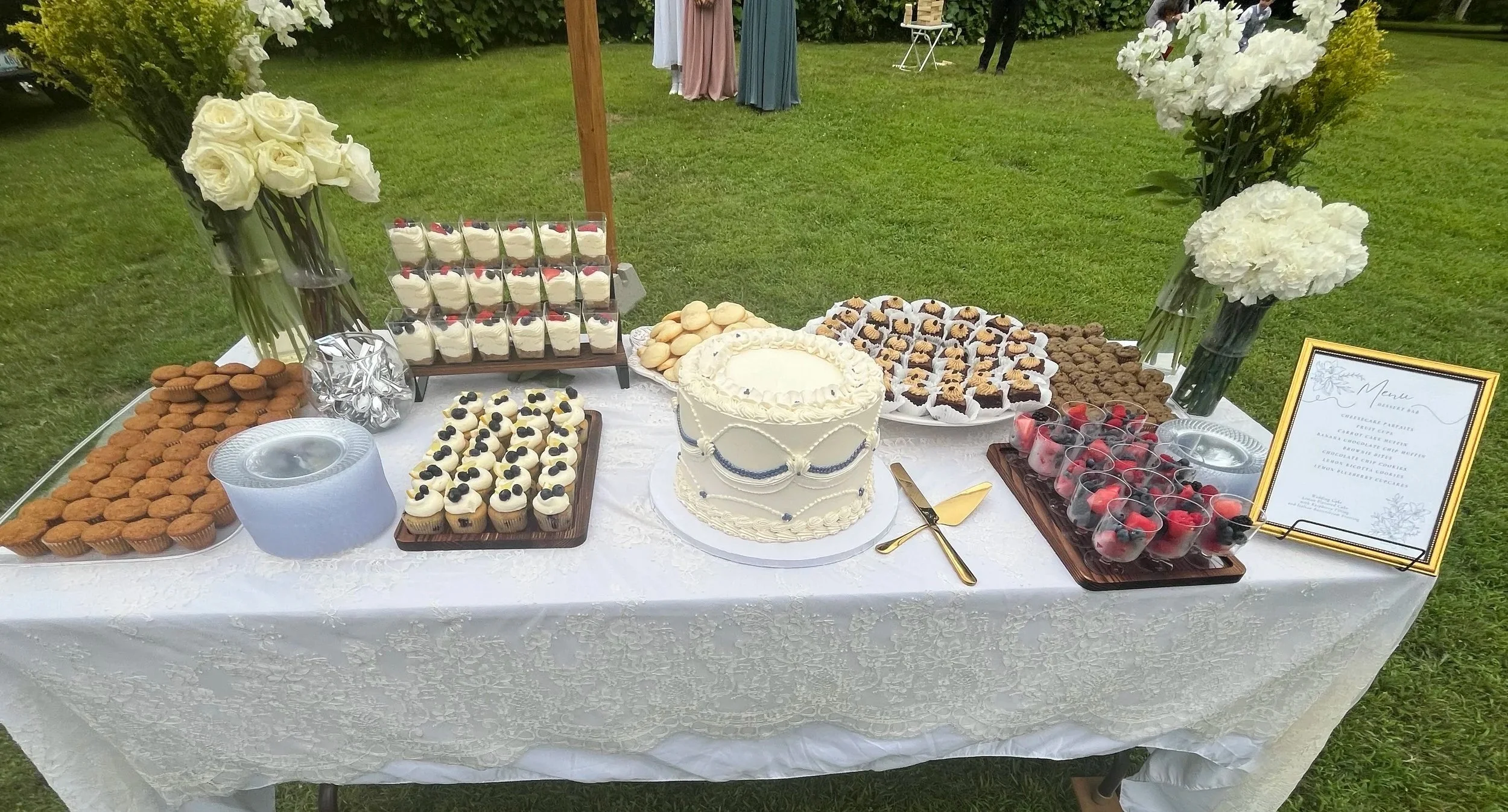 A dessert table with cupcakes, cookies, cake, and parfaits, decorated with flower arrangements and a menu sign in a garden setting.