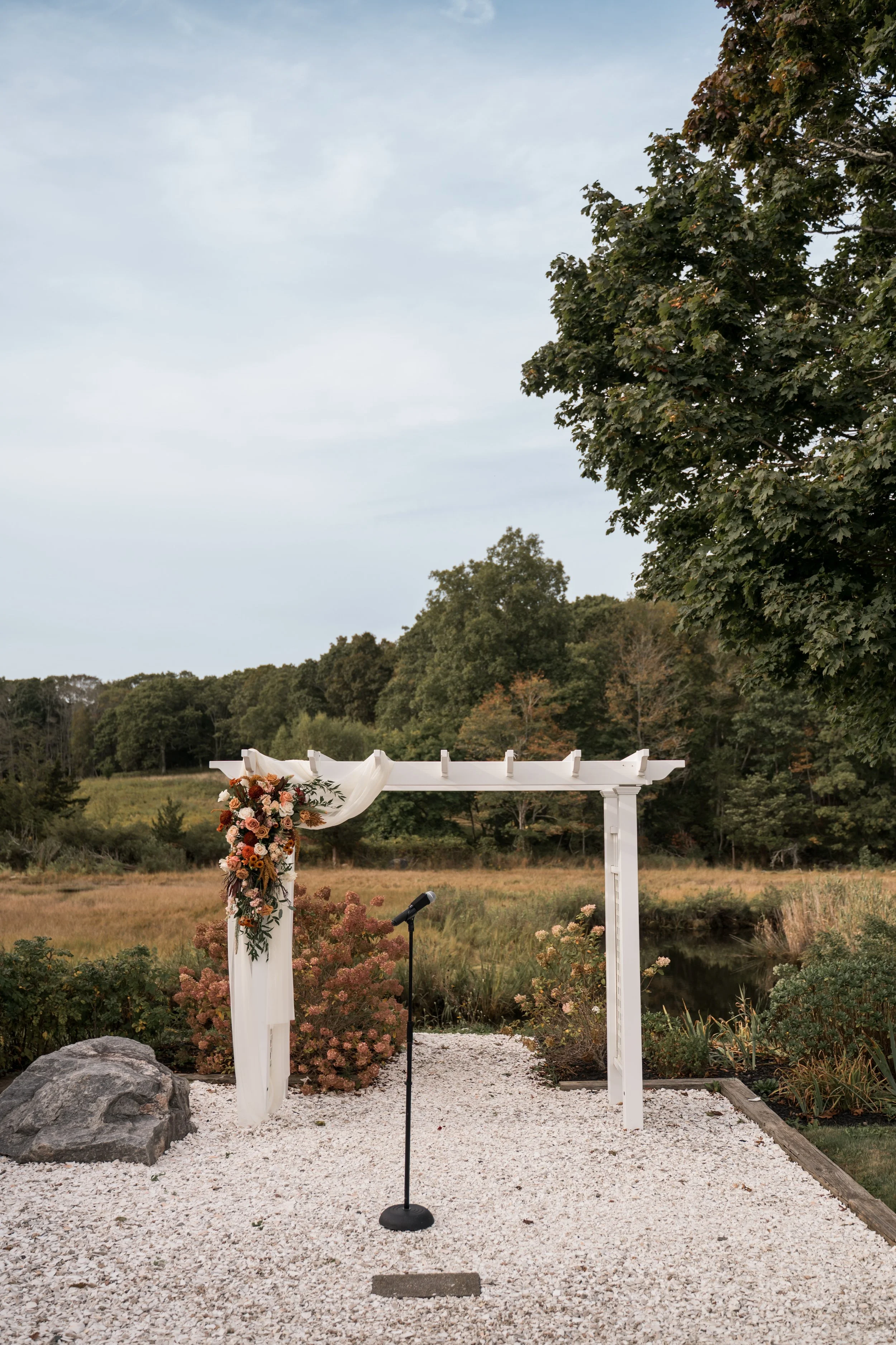 Outdoor wedding arch decorated with flowers and fabric, with a microphone standing in front, set on a gravel path near a pond, surrounded by nature and trees.
