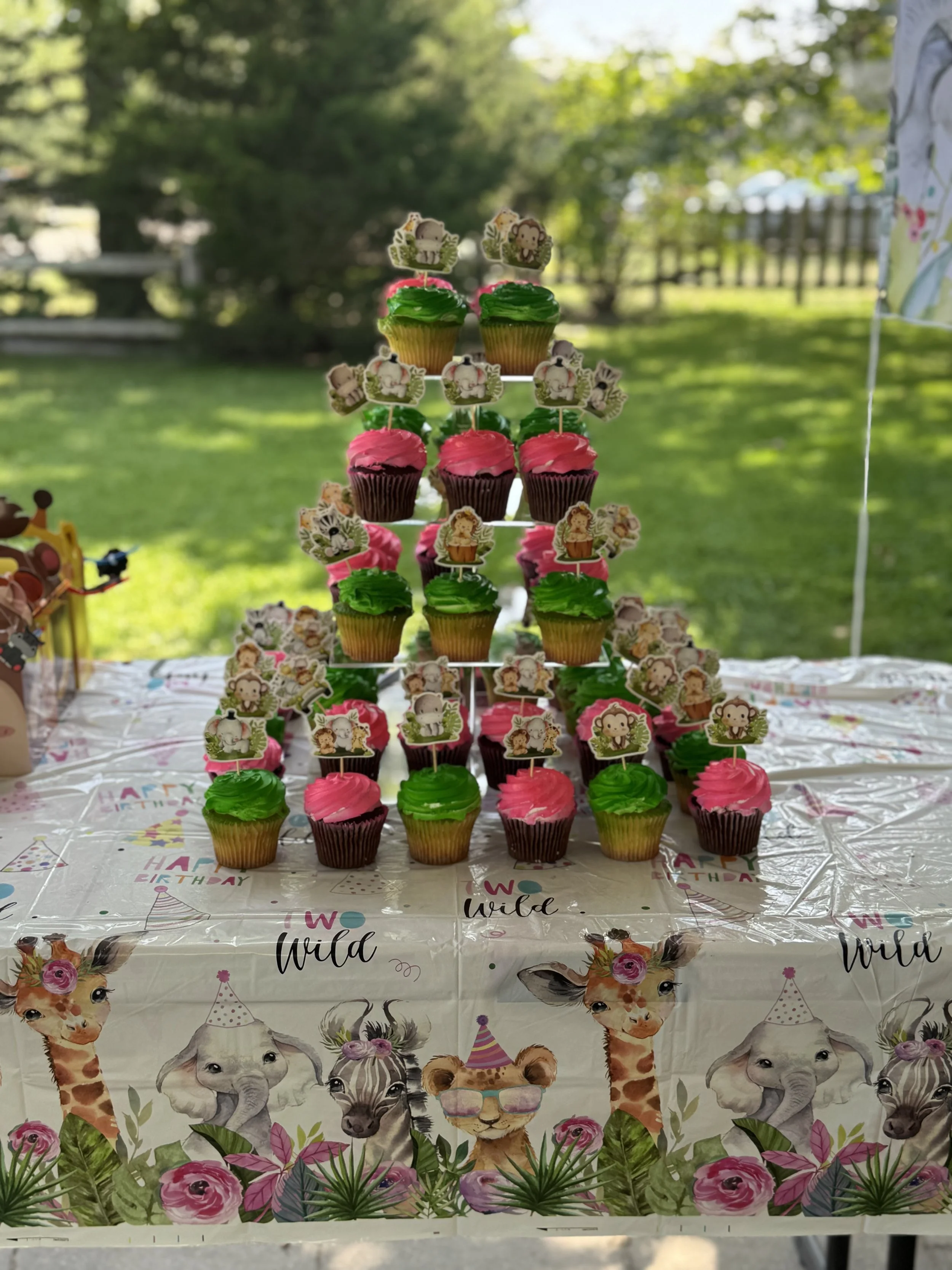 A tiered display of colorful cupcakes with animal themed toppers on a table with a wild animals themed tablecloth, set outdoors with greenery in the background.