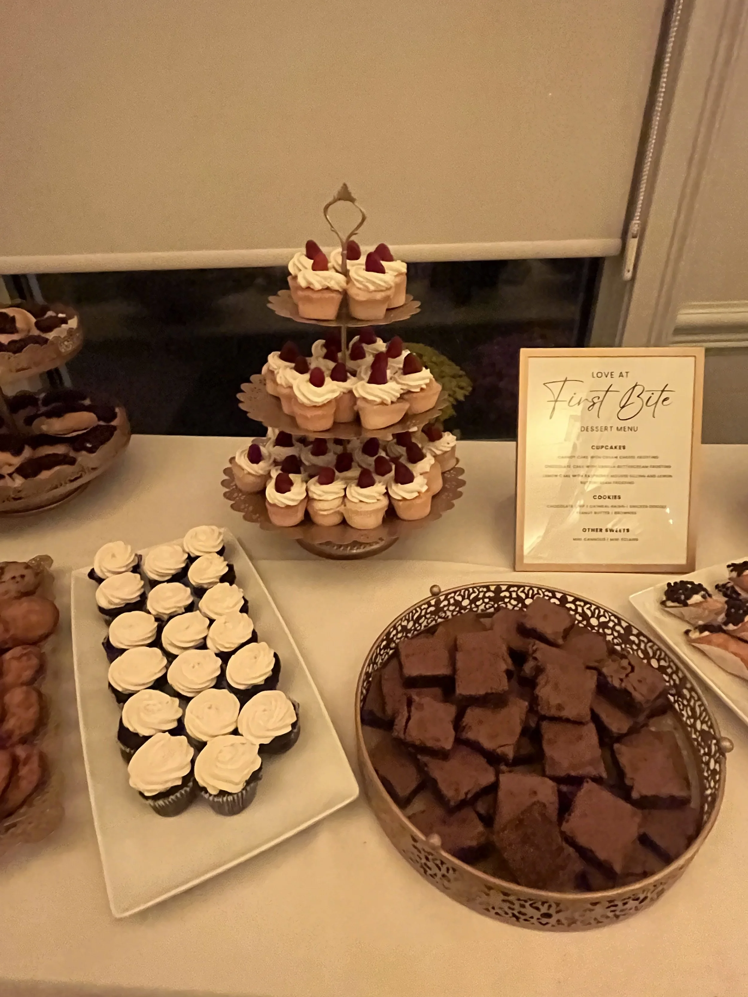 Dessert table with cupcakes topped with raspberries, chocolate brownies, and cupcakes with whipped cream, with a sign that says 'Love at First Bite' and a stand with more cupcakes.