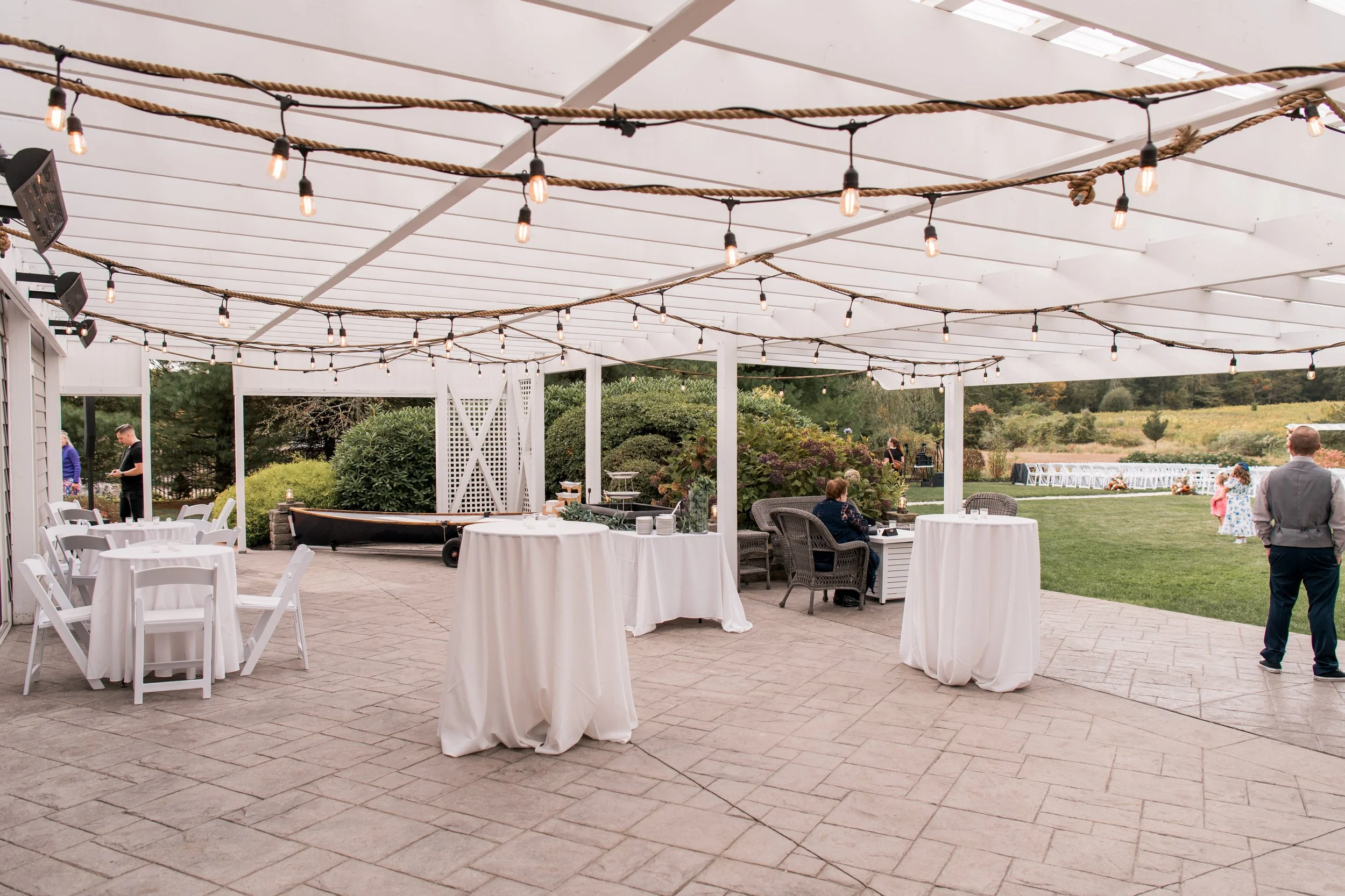 Outdoor cocktail hour with string lights, round cocktail tables with white tablecloths, and a few people in the background. There is greenery and a lawn area outside.