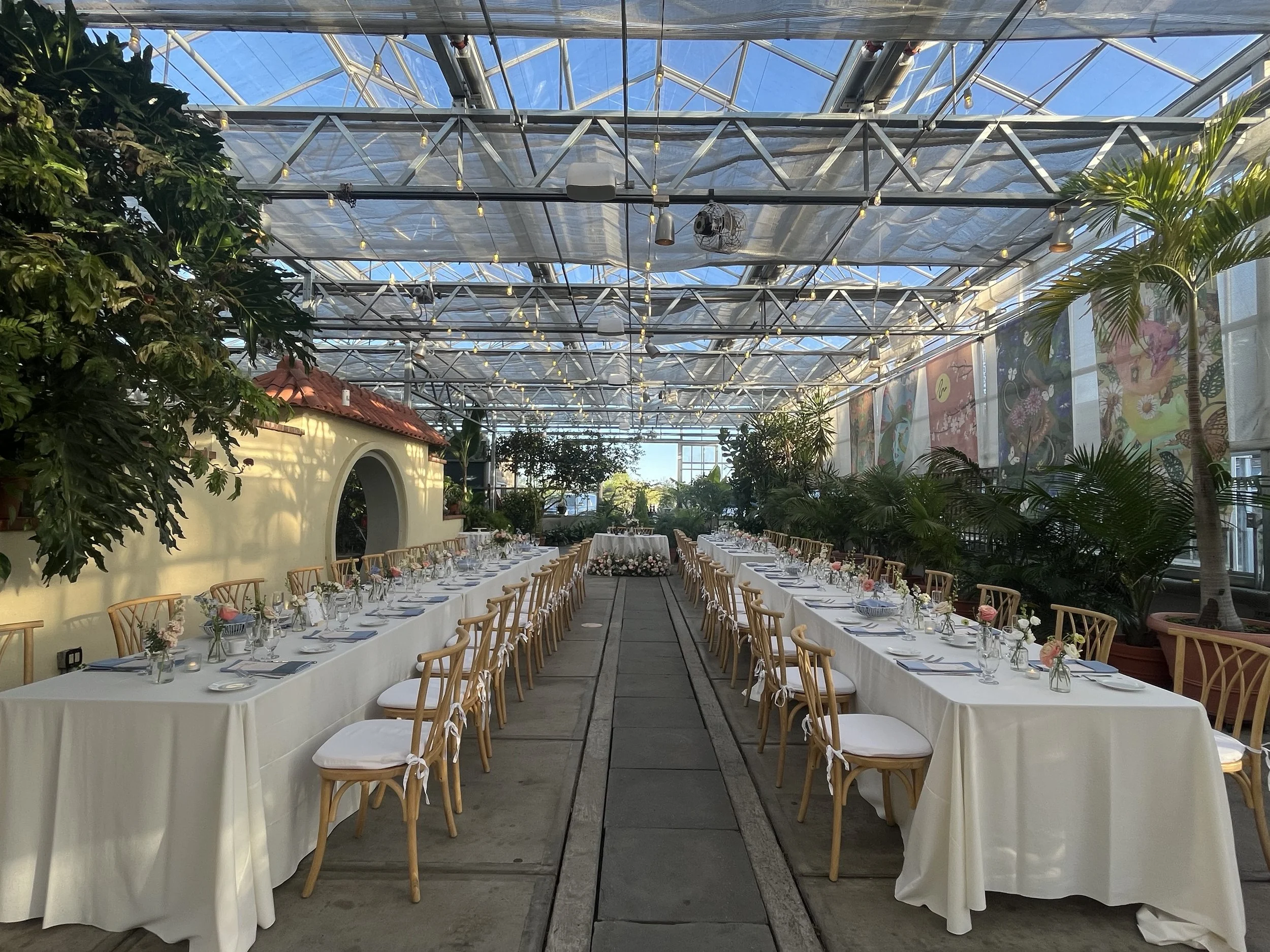 Long banquet tables set with white tablecloths, glassware, and small floral centerpieces inside a bright greenhouse with glass ceiling, lush greenery, and colorful wall art.