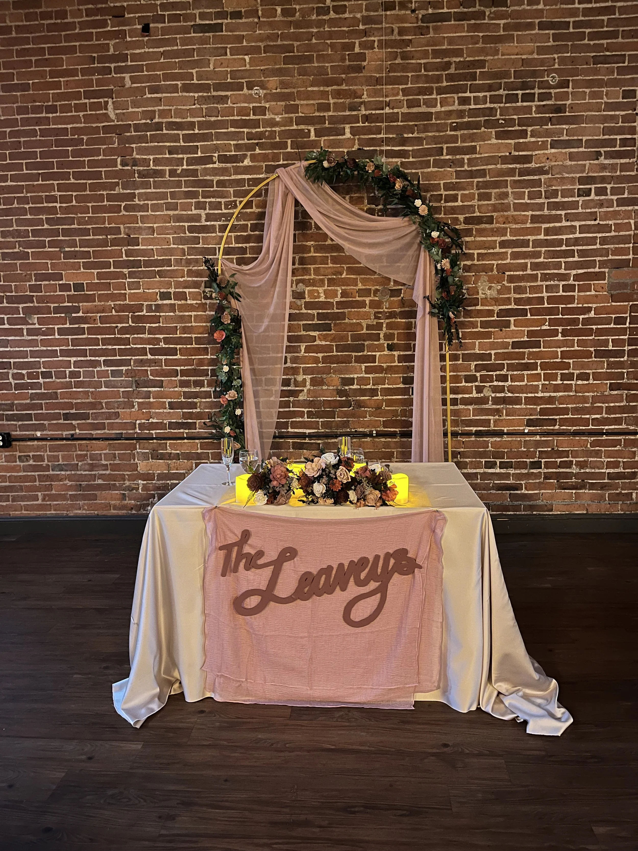 Wedding reception setup with a table in front of a brick wall, decorated with a pink cloth and a floral arrangement, and a backdrop featuring pink drapery, flowers, and a yellow circular frame, with the words "The Laureys" written on the tablecloth.
