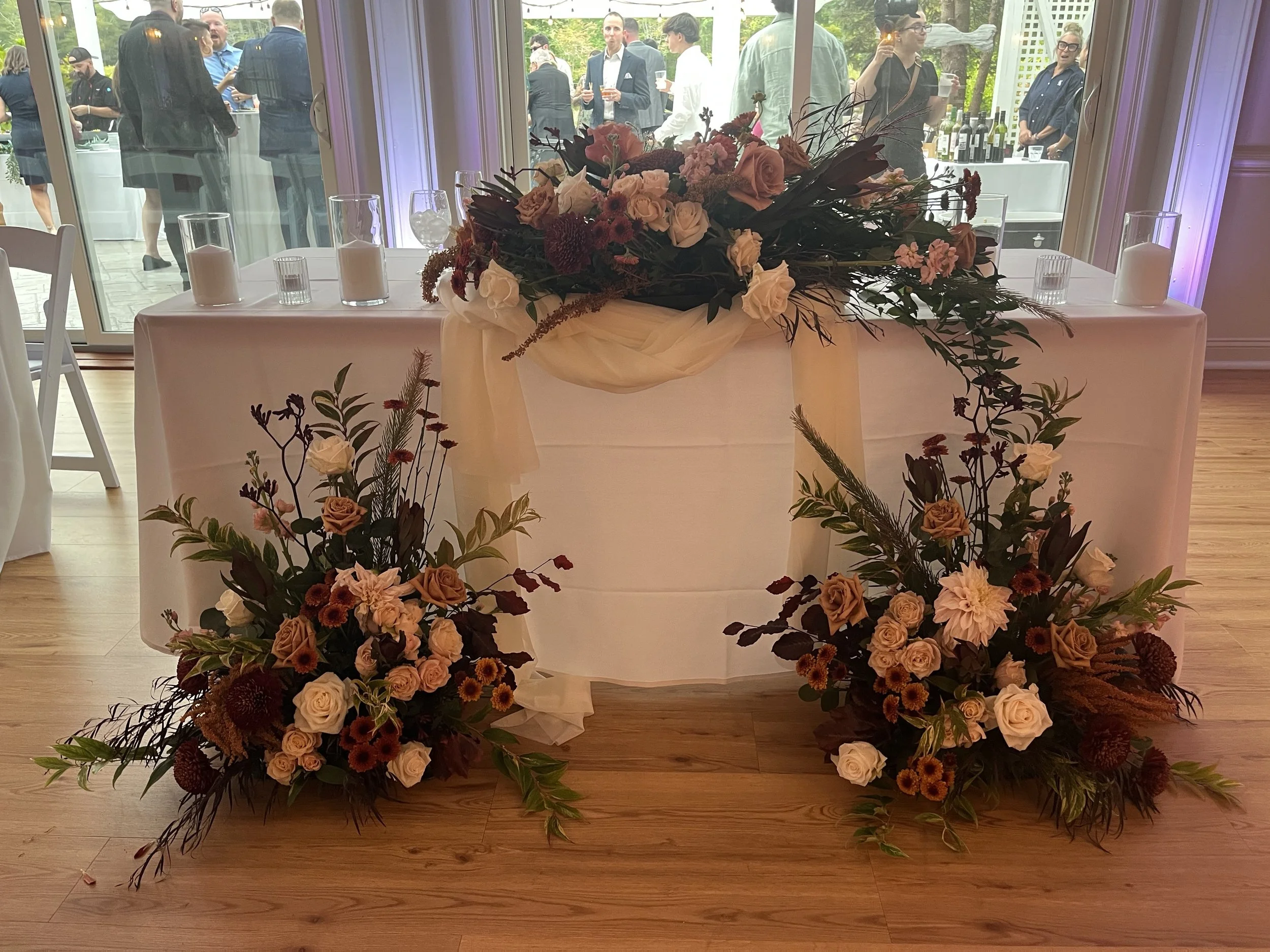 A table decorated sweetheart table with floral arrangements of roses and greenery, set against a backdrop of large windows with guests mingling outside at a social event.