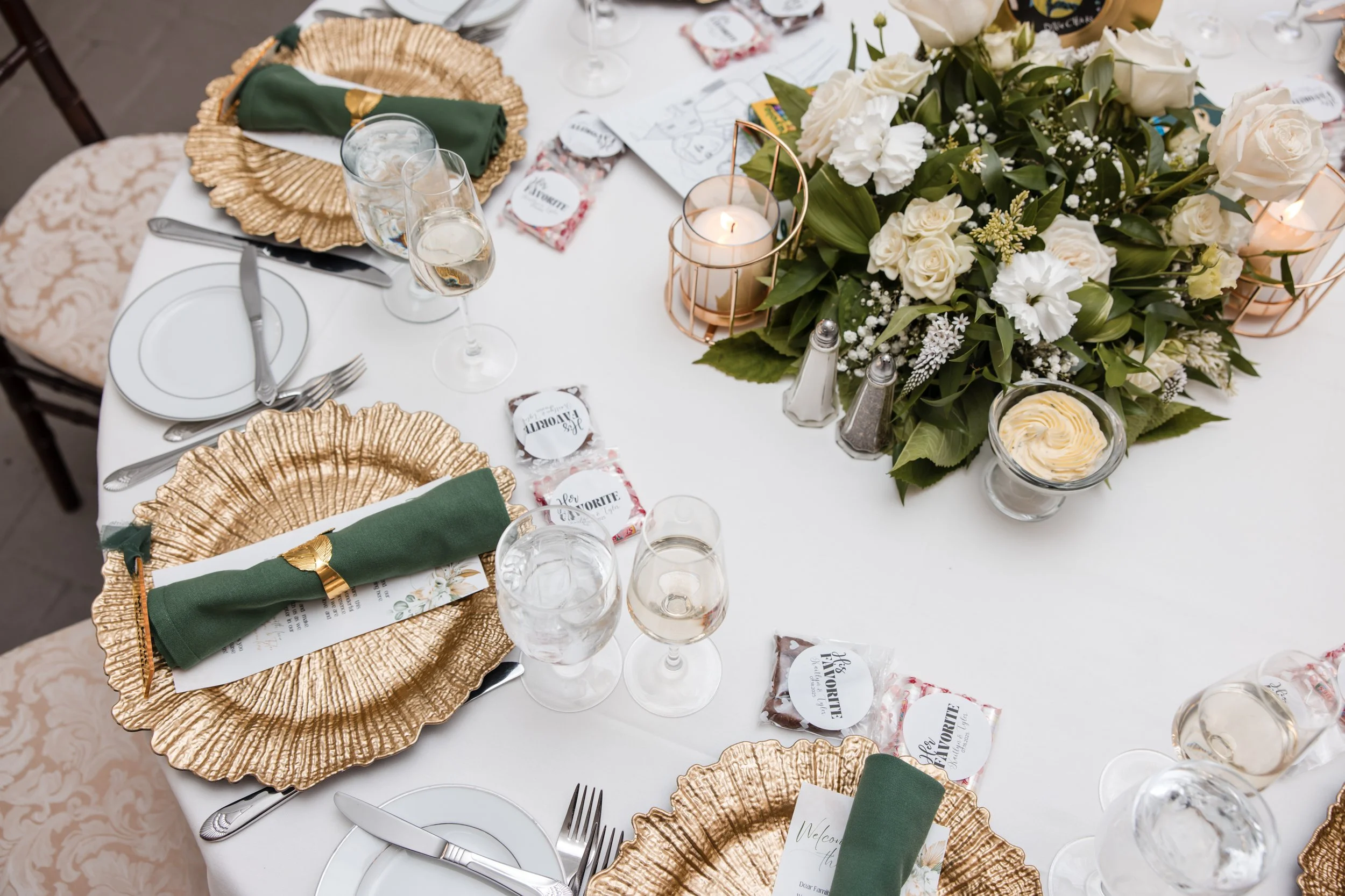Elegant dining table set with gold-rimmed plates, green napkins with gold napkin rings, wine glasses, a floral centerpiece with white roses, lisianthus, and greenery, candles, salt and pepper shakers, and place cards.