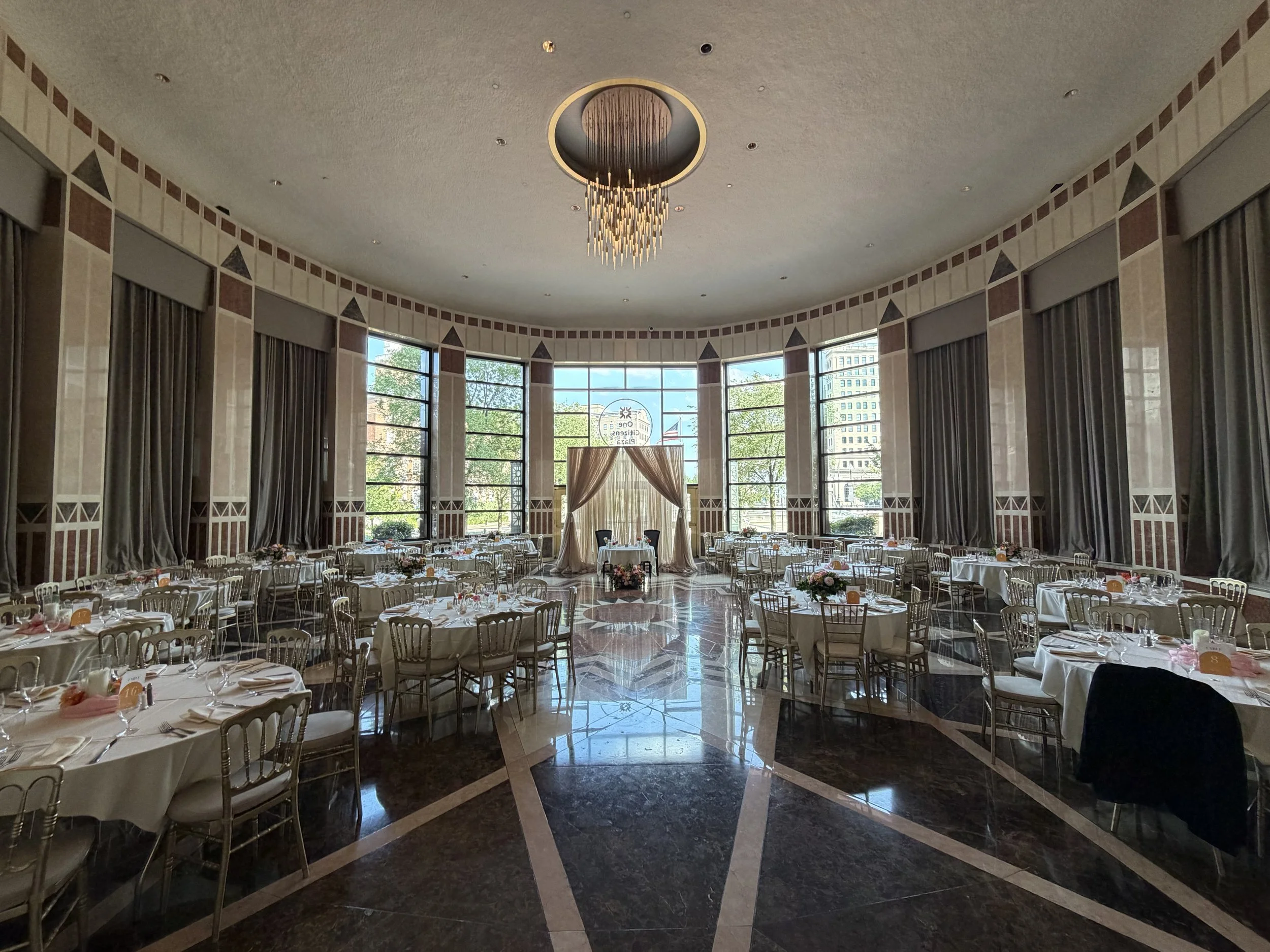 Large banquet hall with round tables set for a wedding or event, decorated with flowers and tableware. Tall windows with drapes and a chandelier hanging from the ceiling. Bright outdoor scenery visible through the windows.