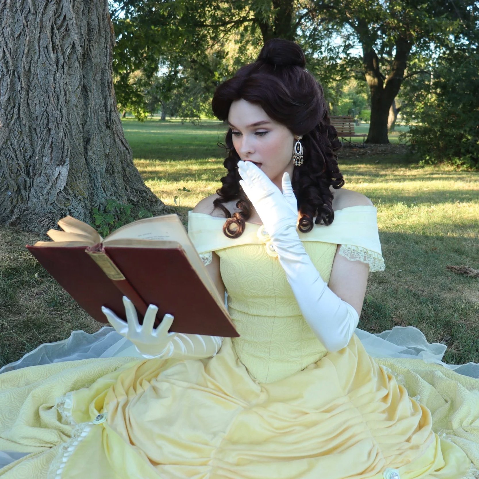 A woman in a yellow ballgown with ivory gloves sits on the grass near a large tree, reading a book.