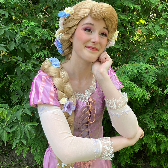 Woman dressed as Rapunzel, with long blonde braid decorated with flowers, wearing a pink and cream dress with lace details, standing outdoors among green foliage.