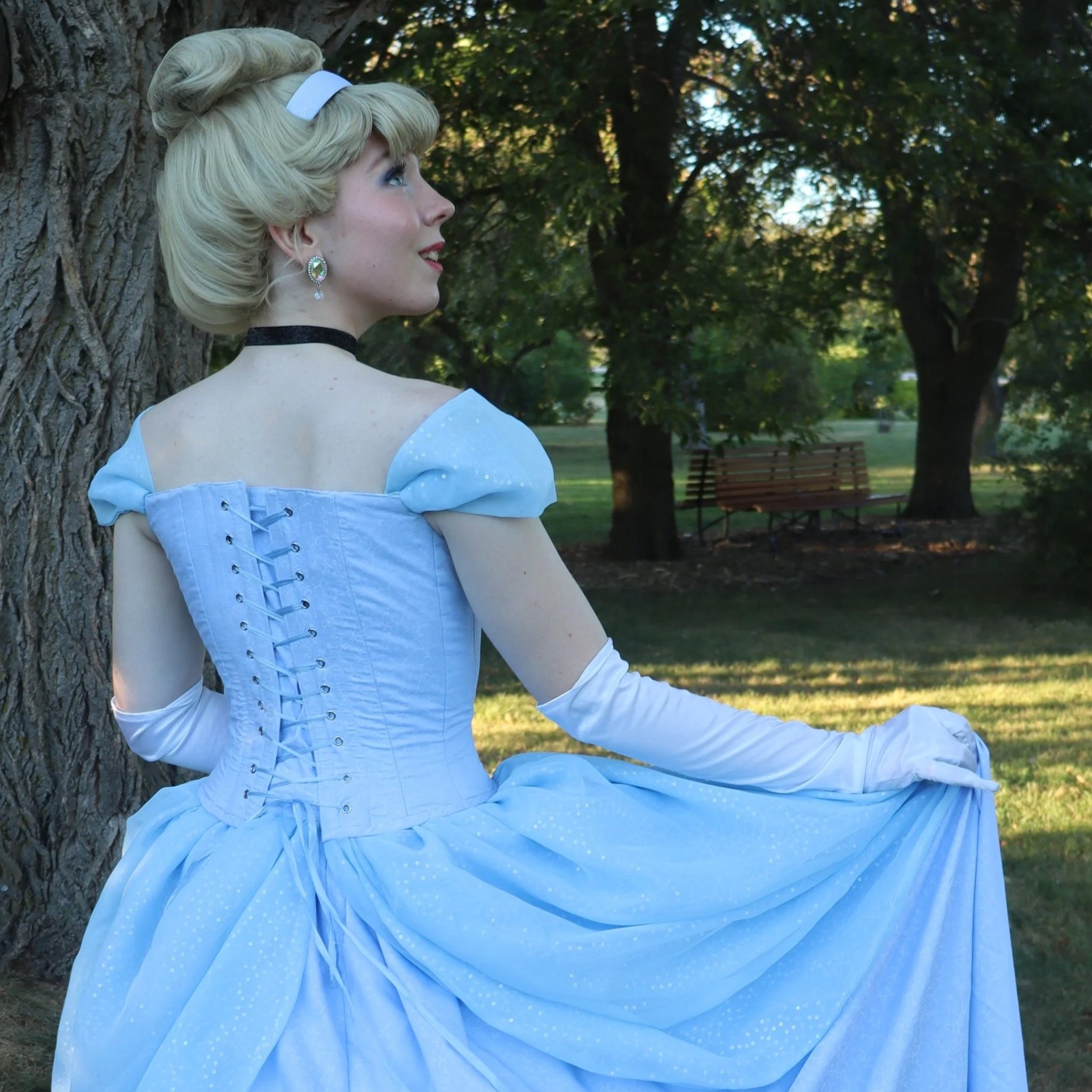 A woman dressed as Cinderella sitting against a tree in a park, wearing a light blue gown with white gloves and looking to the side.