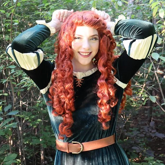 A woman with long, curly red hair smiling in a forest setting, wearing a dark green and beige dress with a brown belt, holding her hands above her head.