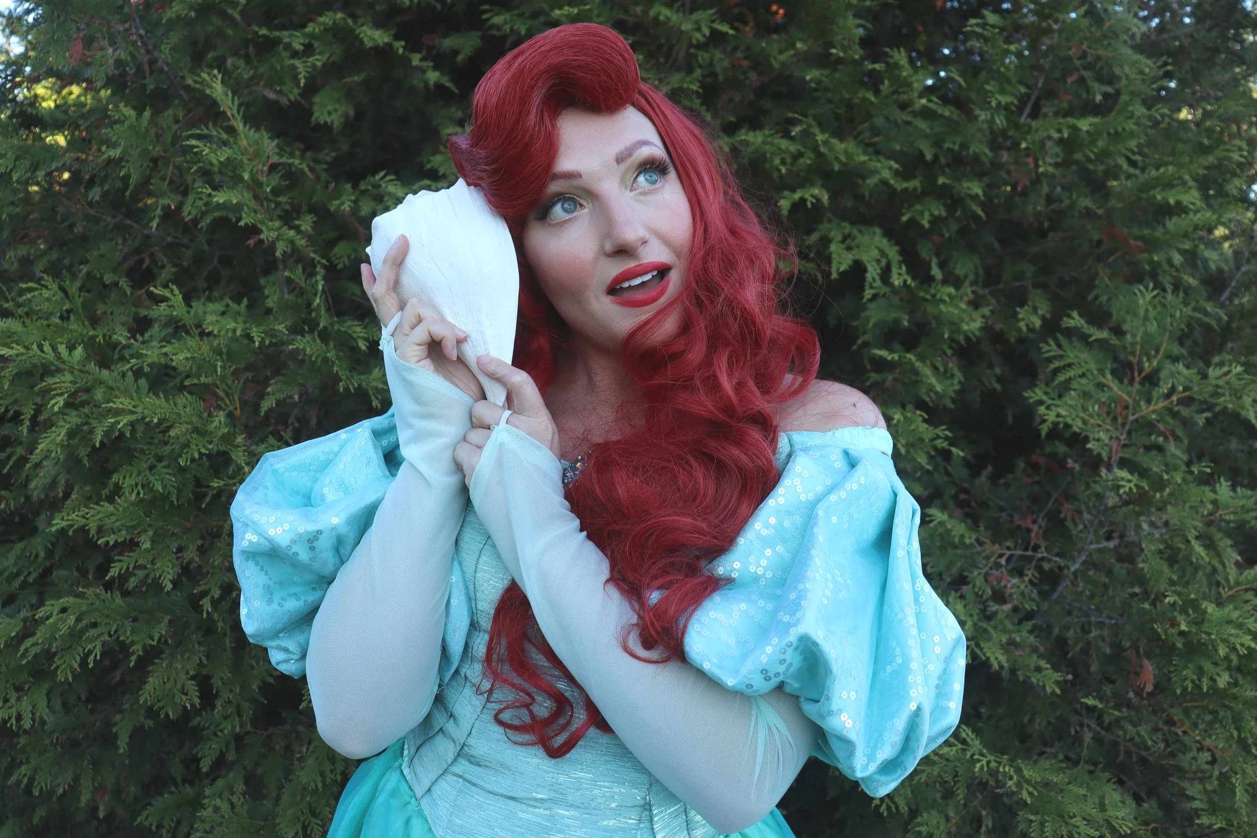 A woman with long red curly hair, wearing a light mint dress with puffed sleeves, is holding a seashell to her ear and looking upwards with a dreamy expression, standing outdoors in front of greenery.