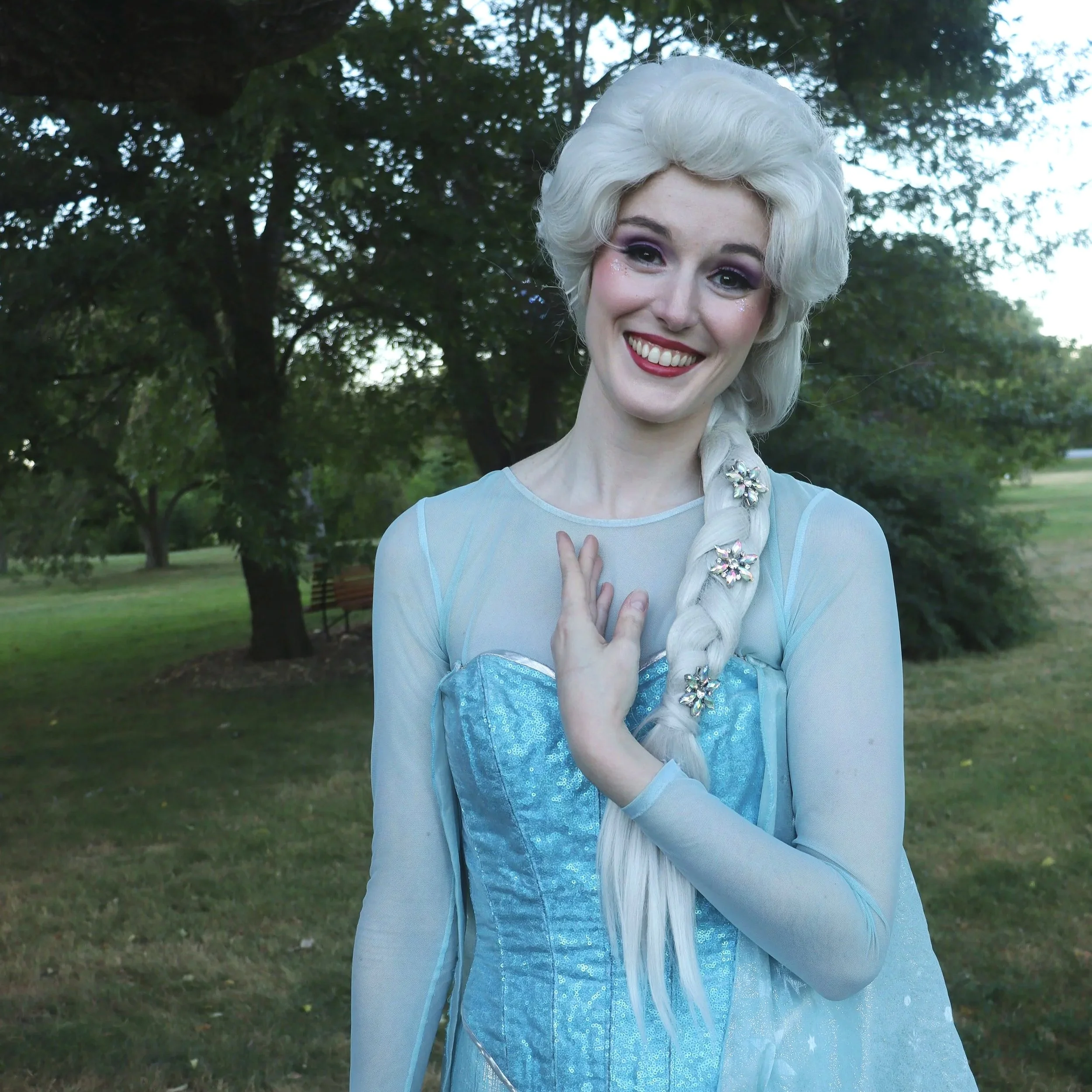 A woman dressed as a fairy tale princess with long, platinum blonde hair wearing a light blue dress and sheer long sleeves, standing outdoors in a park with trees and grass, smiling.