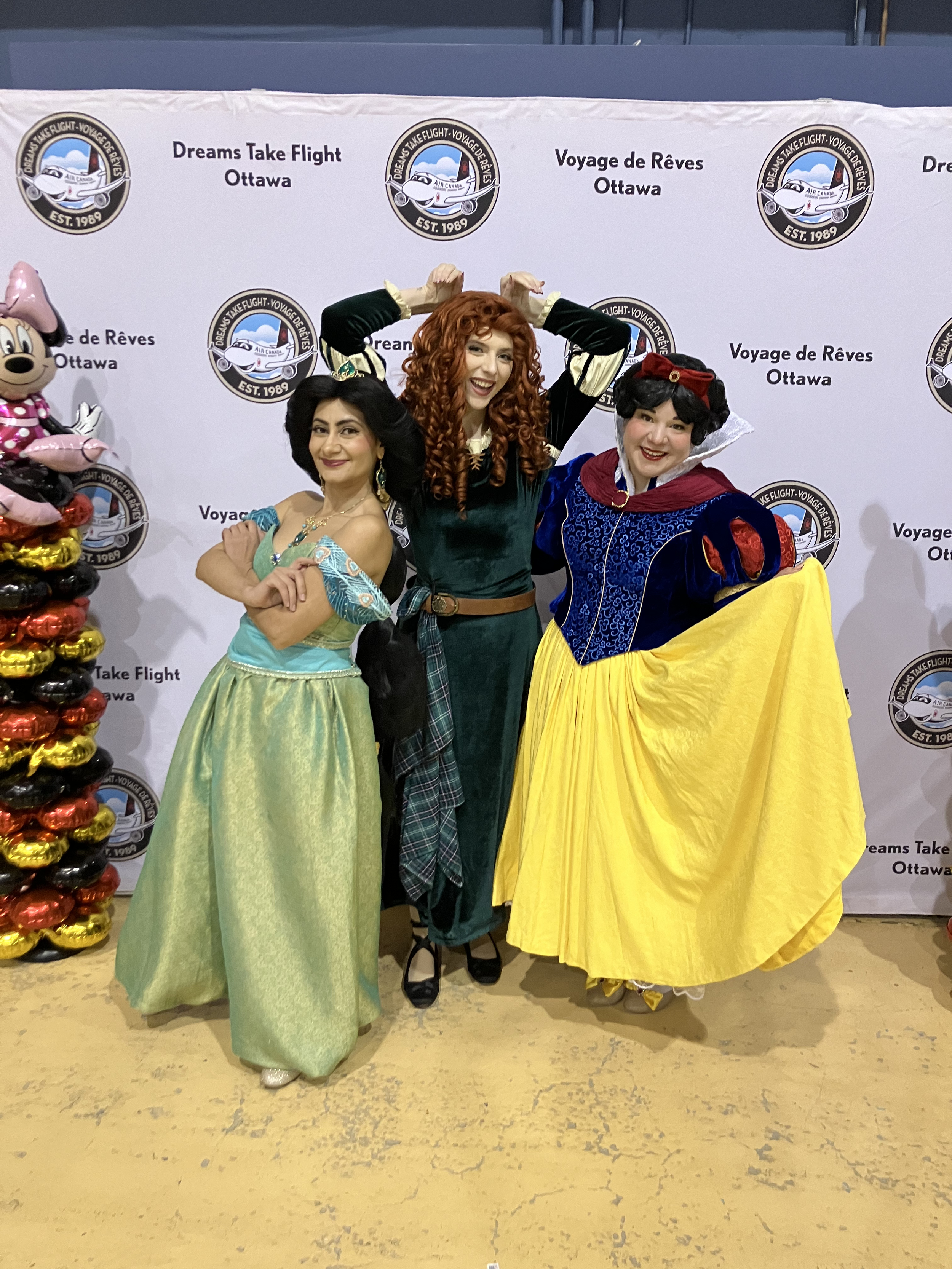 Three women dressed as princesses posing in front of a backdrop with logos for Dreams Take Flight Ottawa, with Minnie Mouse balloons on the left.