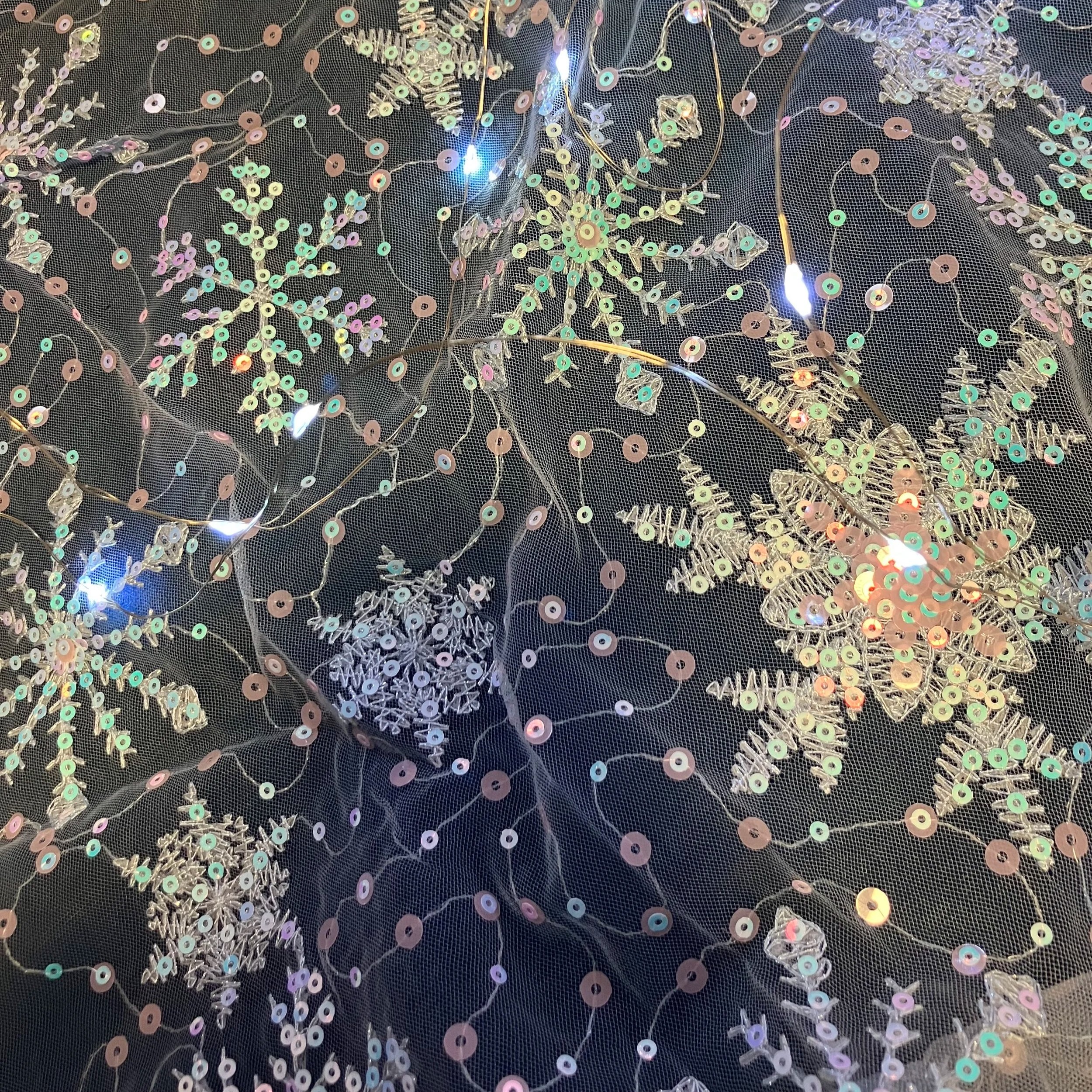 Close-up of a black fabric with iridescent sequins and embroidered snowflake patterns, accented with small LED lights.