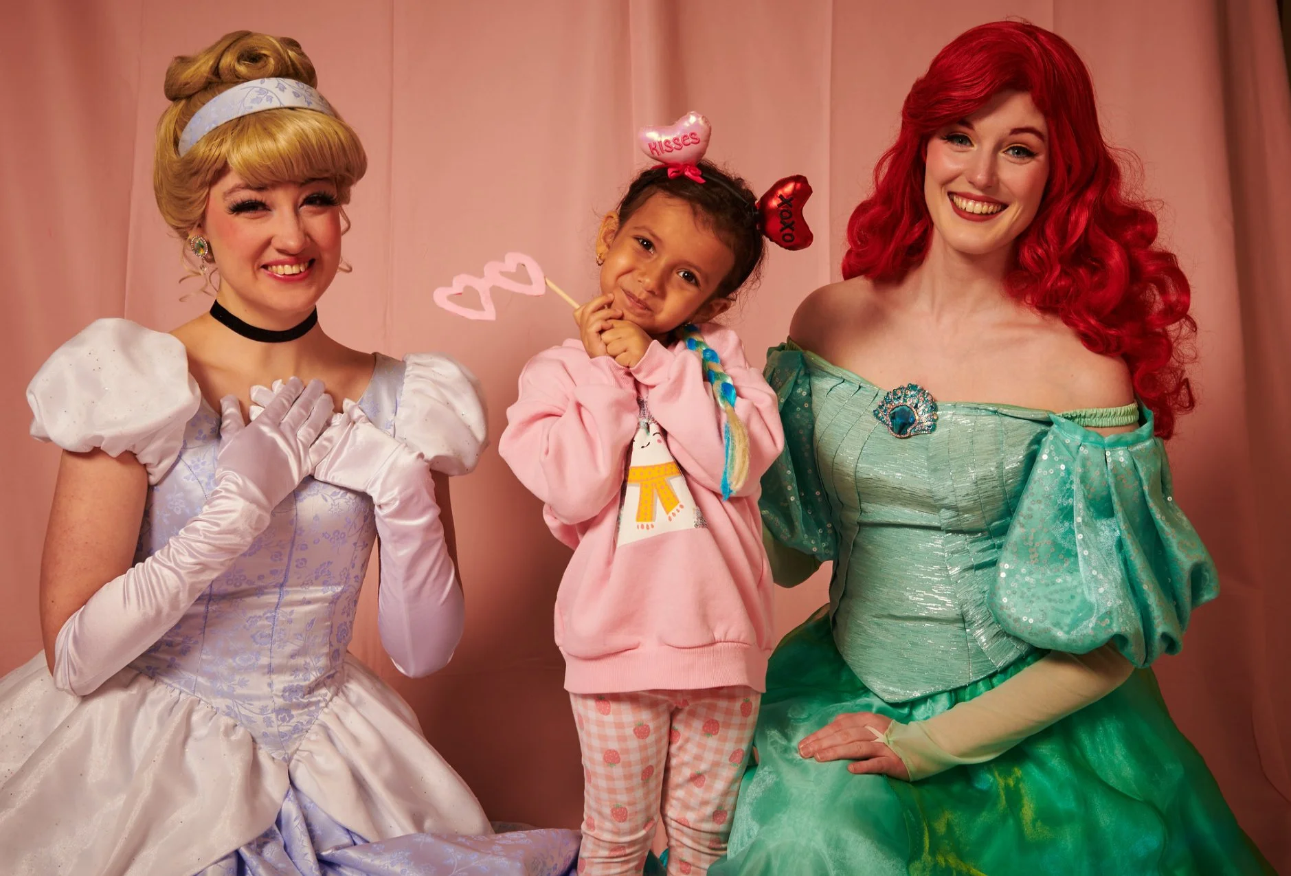 A young girl dressed in a pink sweatshirt holding heart-shaped Valentine decorations, with two women in princess costumes, one of whom is from The Little Mermaid. The girl is standing between the women, smiling, against a pink backdrop.