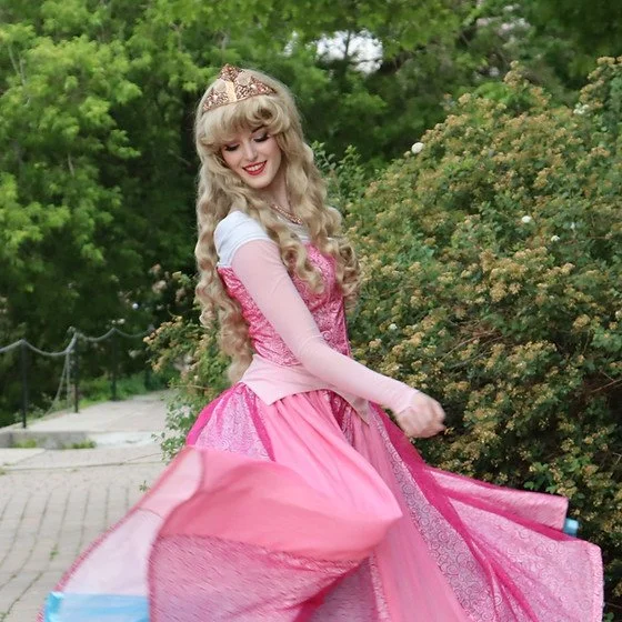 A woman in a pink princess dress and tiara twirling outdoors with greenery in the background.