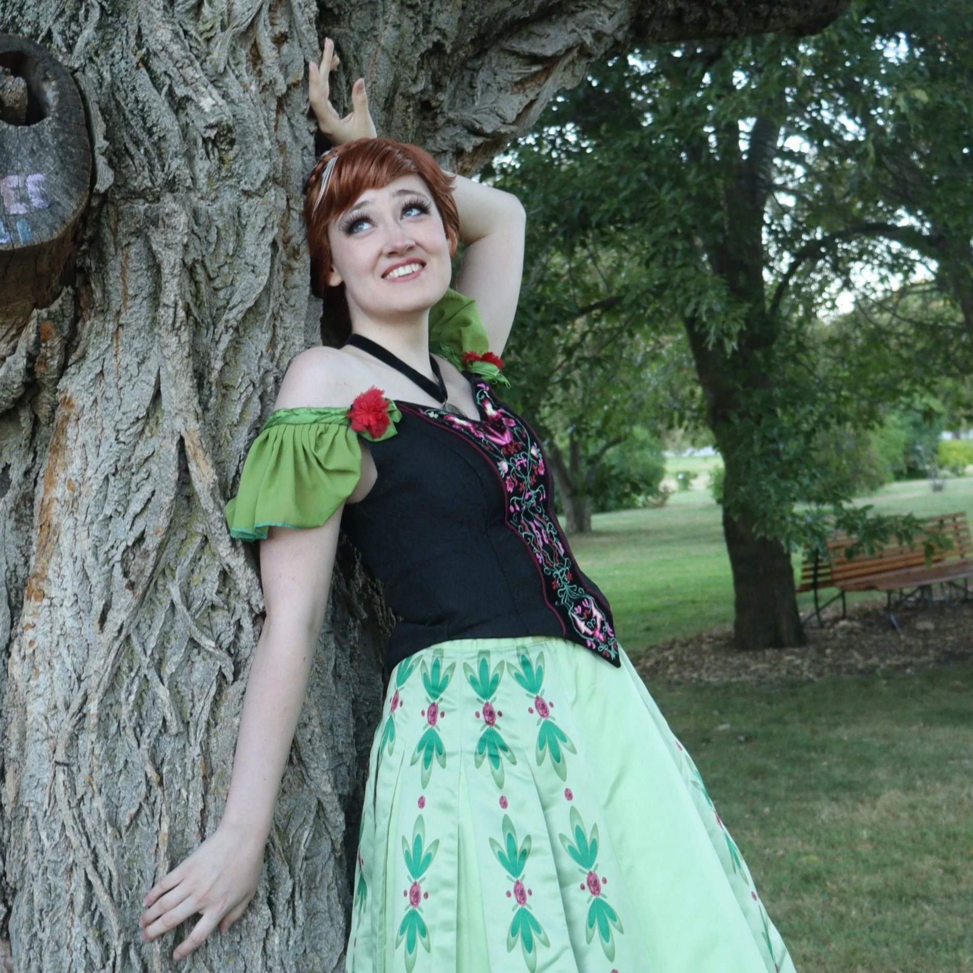 A woman with red hair in an updo and a black top with pink embroidery, wearing a green skirt with green and pink floral patterns, leaning against a large tree with her head tilted back and smiling in a park.