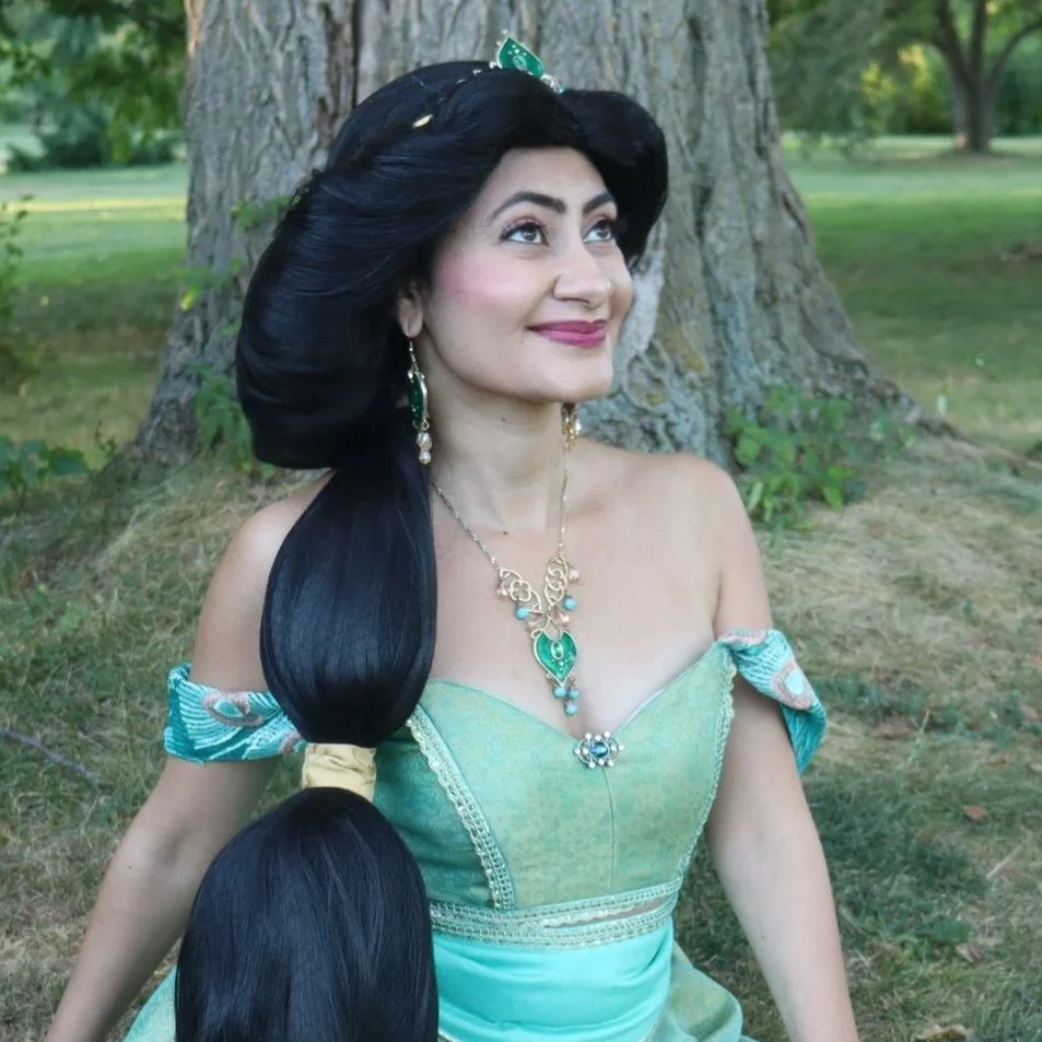 A woman dressed in an off-shoulder green gown with jewelry, sitting outdoors near a tree, with a joyful expression.