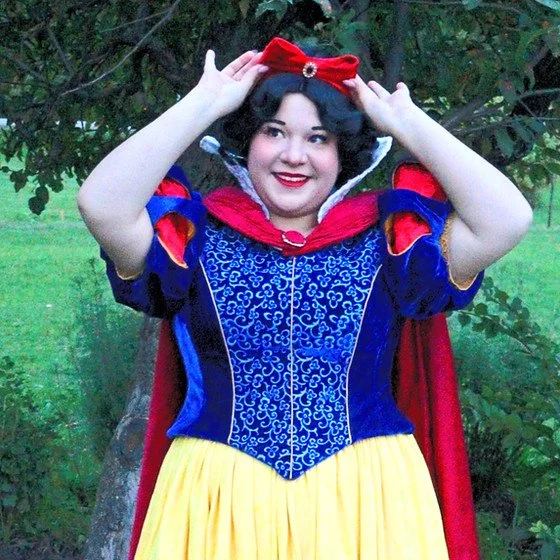 Person dressed as Snow White adjusting a red bow headband outdoors with green foliage in the background.