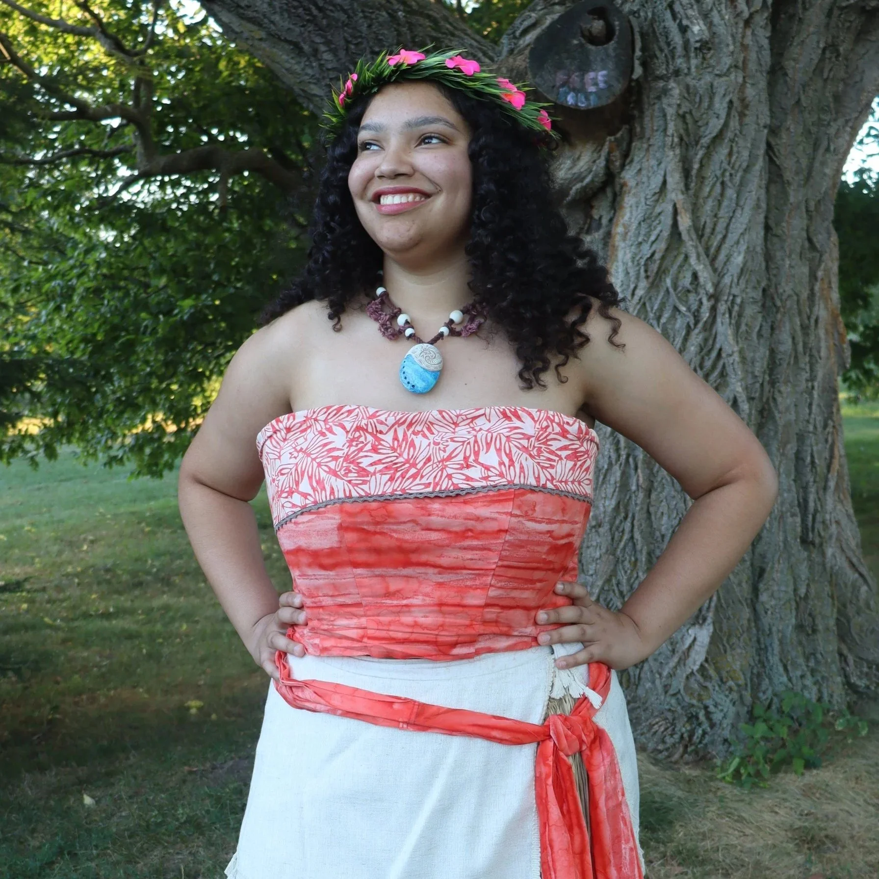 A woman with curly black hair, wearing a flower crown, smiling and standing outdoors next to a large tree. She is dressed in a strapless top with a red and white pattern and a white skirt with an orange sash.