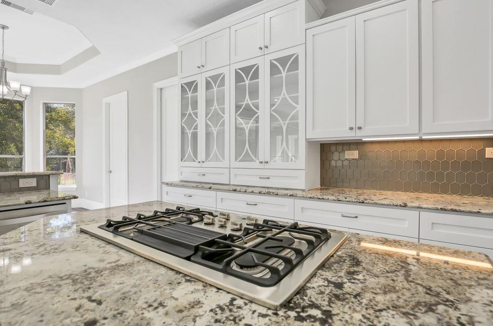 Modern kitchen with granite countertops, gas stove, white cabinets, glass-front cabinet doors, and a hexagon tile backsplash. Large windows visible in the background.