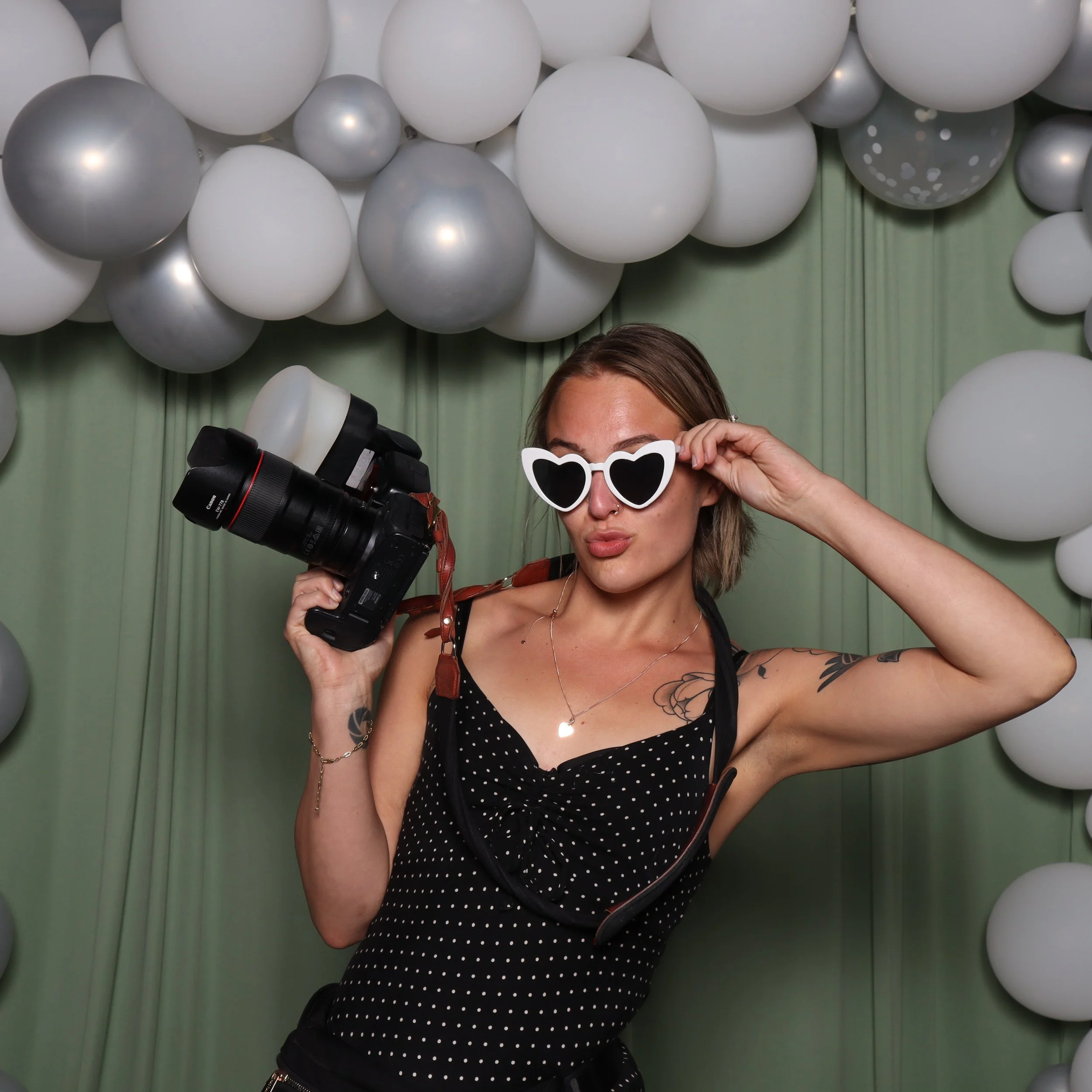 A woman with tattoos wearing a polka-dot top and white heart-shaped sunglasses posing with a camera in front of a green curtain decorated with white and silver balloons.