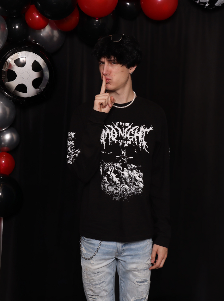 Young man with black curly hair, wearing a black band T-shirt, ripped jeans, a pearl necklace, and sunglasses on his head, standing in front of a black background decorated with black, red, and silver balloons, some of which are shaped like wheels.
