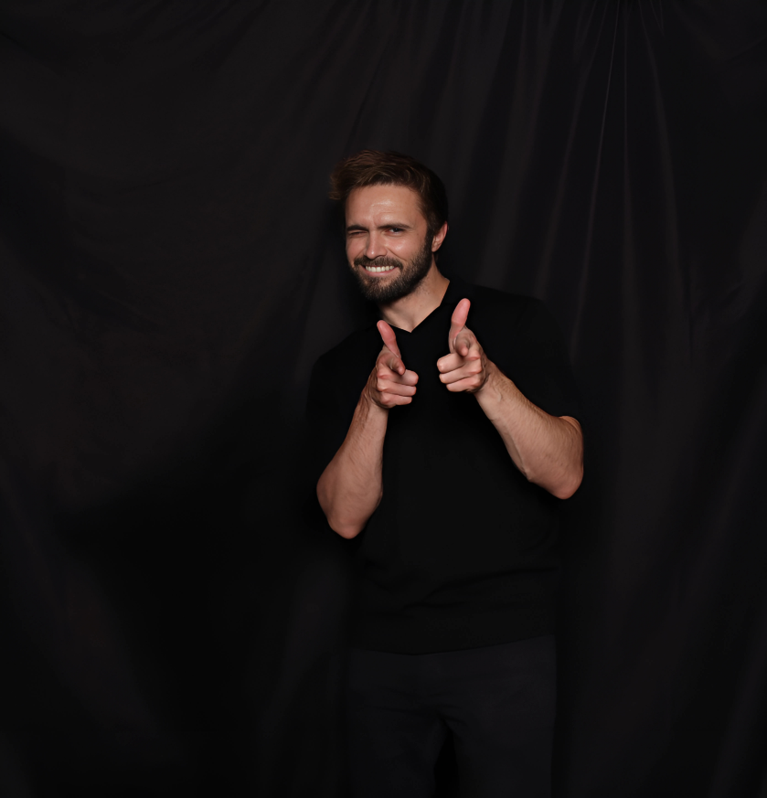 A smiling man with a beard, wearing a black shirt, pointing both index fingers forward, against a dark backdrop.