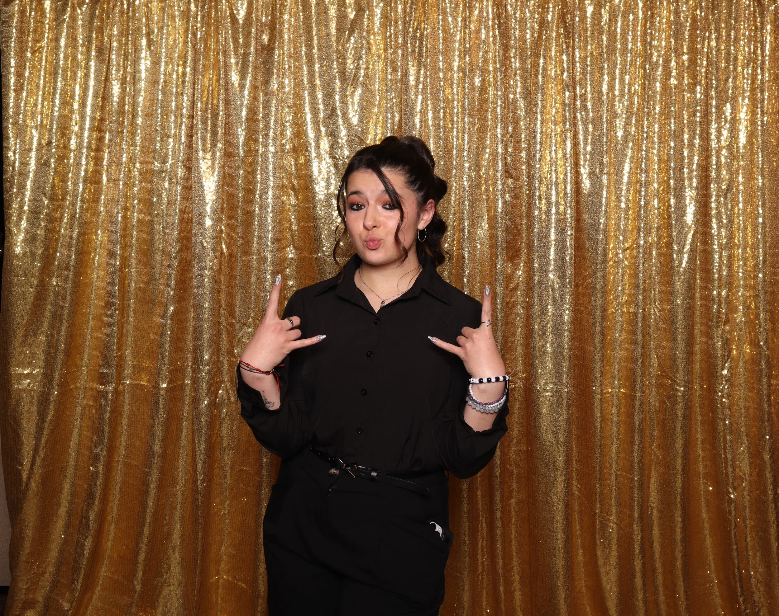 Young woman with dark hair styled in loose curls, making a playful face and rock hand gestures, in front of a shiny gold sequin curtain.
