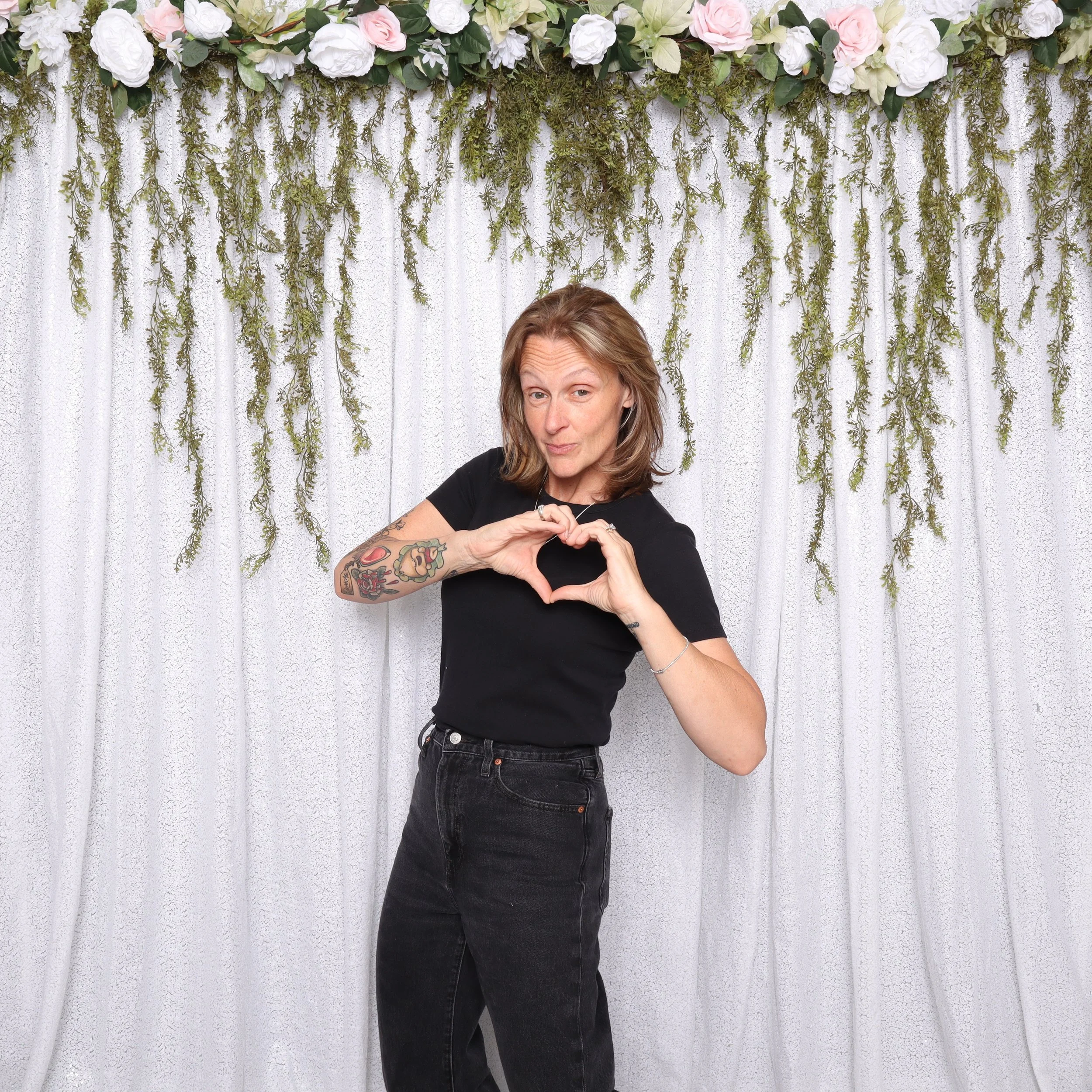 A woman with tattoos on her right arm making a heart shape with her hands in front of a white curtain decorated with hanging greenery and white and pink flowers at the top.