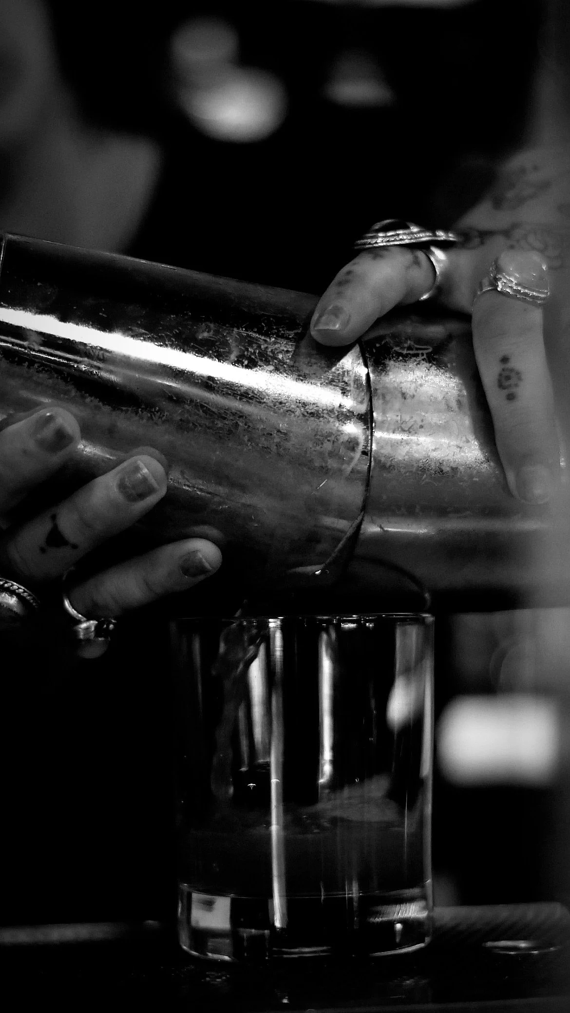 Black and white photo of hands pouring a drink from a metal shaker into a glass.