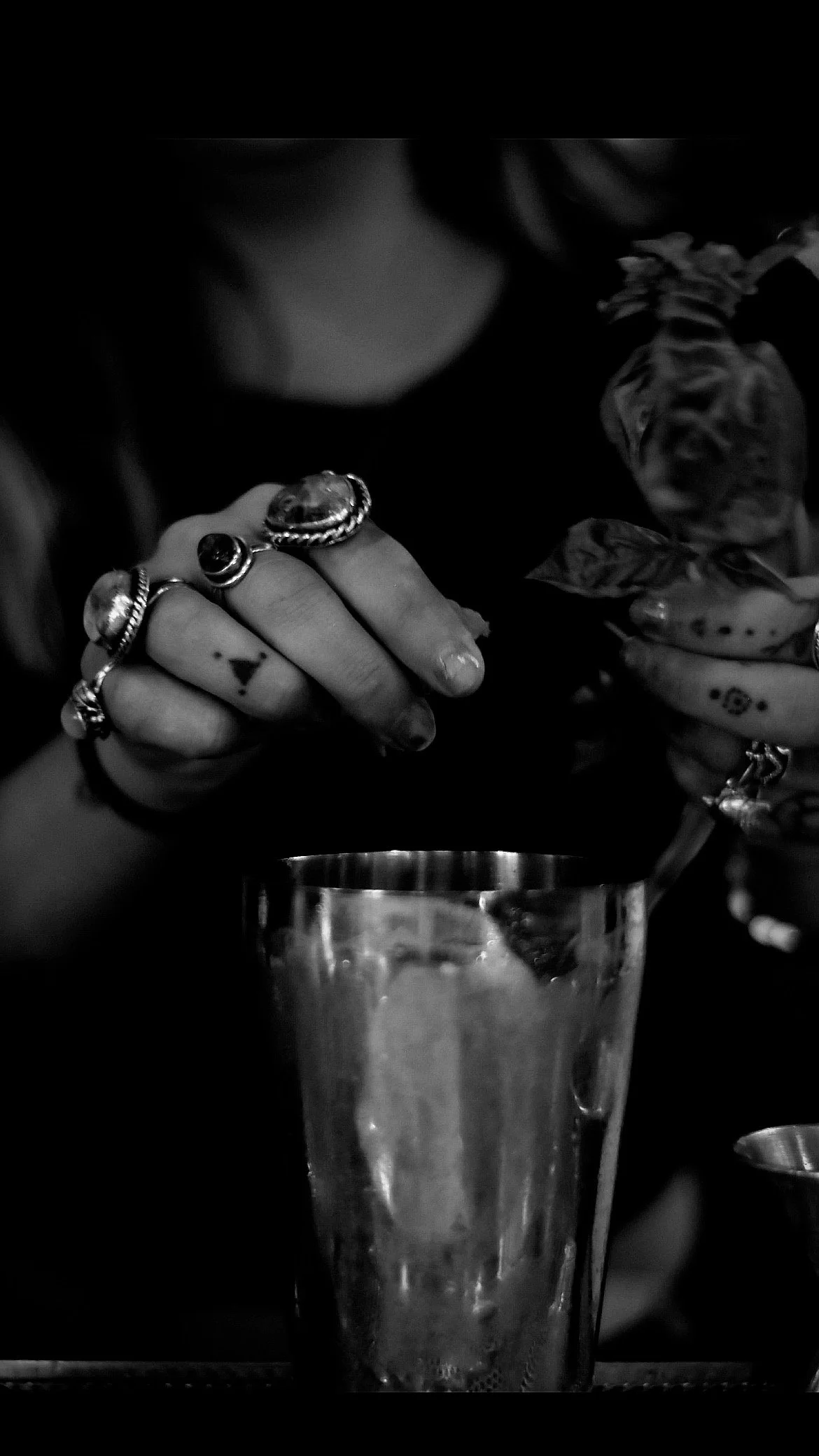 A person with multiple rings on their fingers is squeezing a lemon slice over a tall glass of drink with ice. The person has tattoos on their fingers and is holding the lemon with one hand while using the other hand to squeeze.