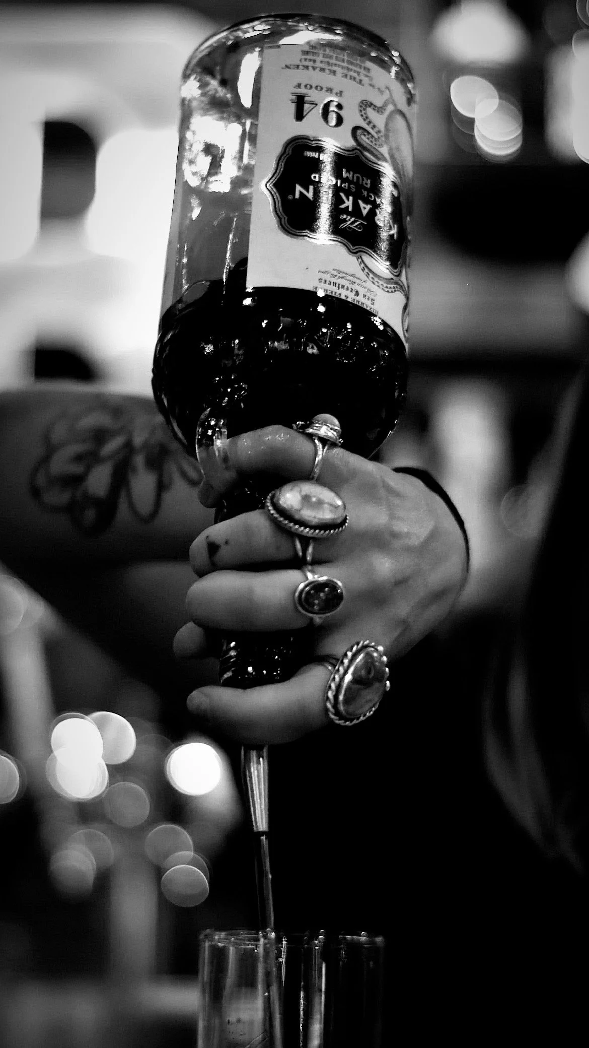 Black and white photo of a person’s hand, adorned with multiple large rings, holding a wine glass with a bottle of red wine. The wine is pouring into the glass from the bottle. The background is blurred with bokeh lights.