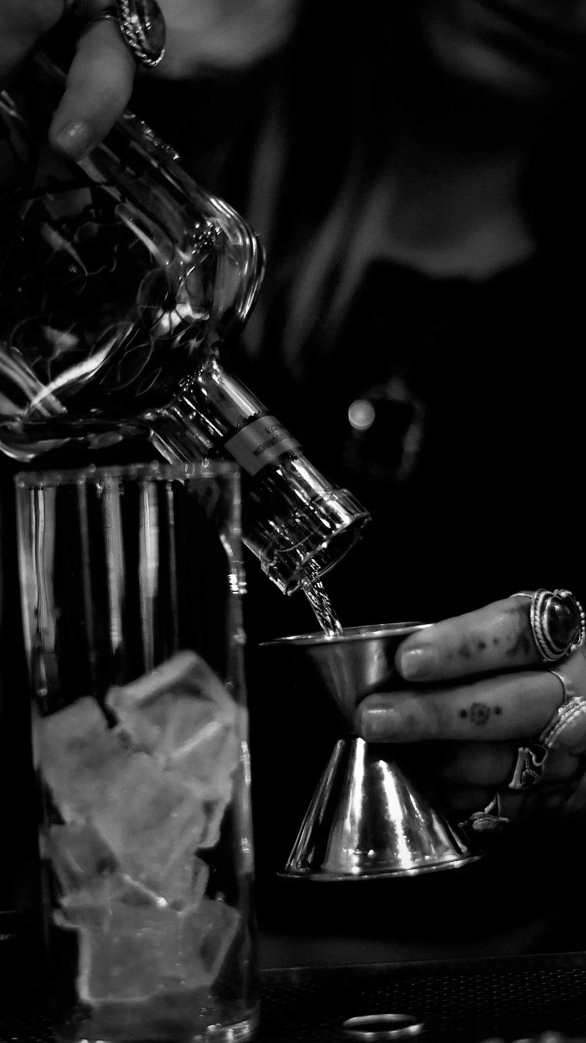 Black and white photo of a person pouring a clear drink into a metal shaker, with a glass of ice nearby.