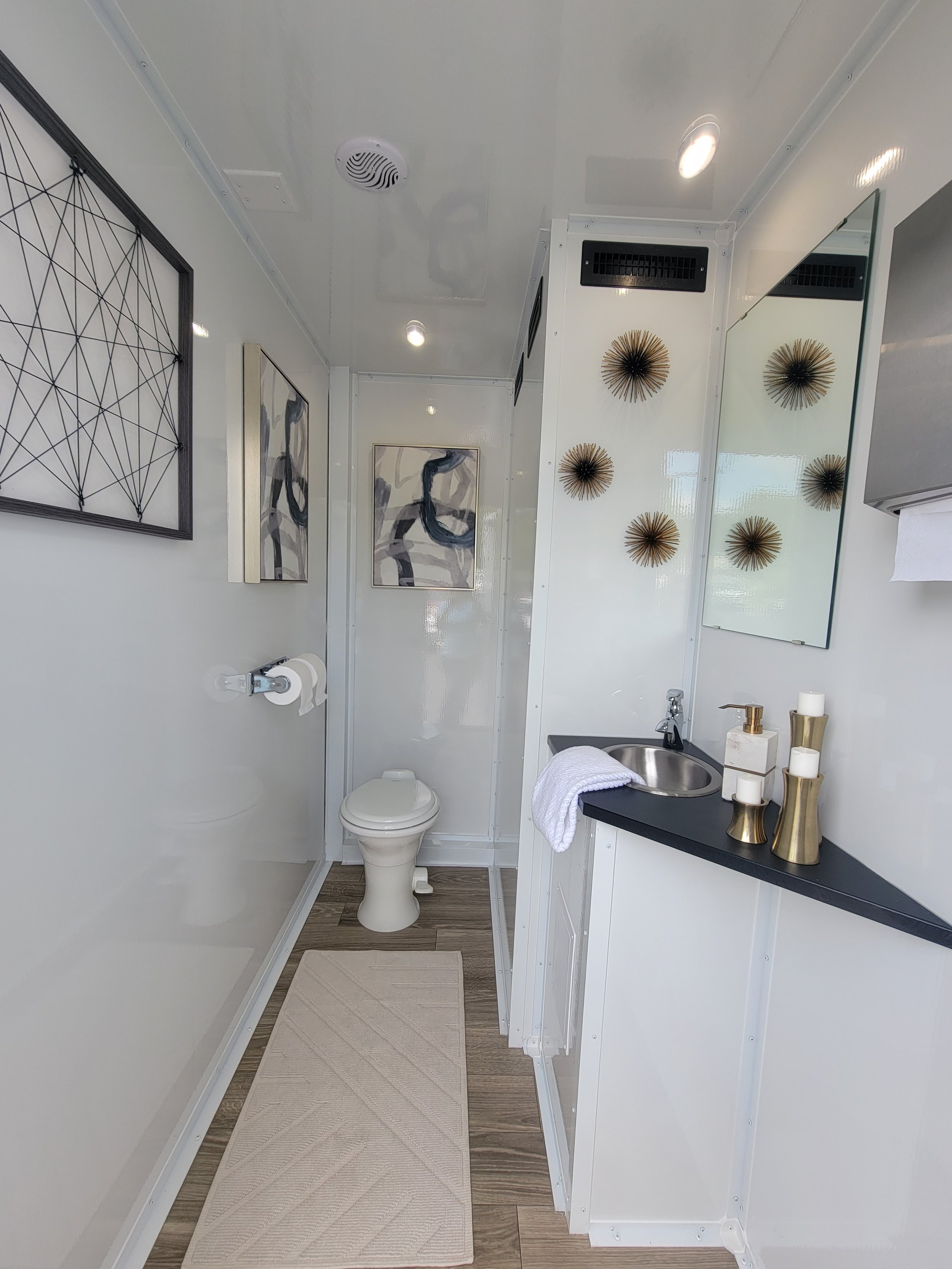 Modern, white bathroom with a toilet, sink, and decorative elements such as wall art and sculptures. Features include a white rug, wall-mounted toilet paper holder, and golden accents like candle holders on the sink counter.