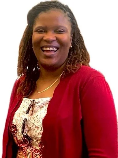 A smiling woman with dreadlocks wearing a red blazer over a patterned blouse and a gold necklace.