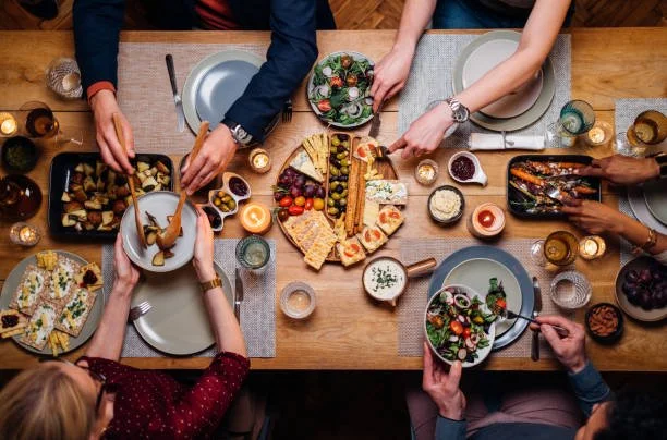 A family gathered around a wooden dinner table sharing a meal with various dishes, salads, and desserts, seen from above.