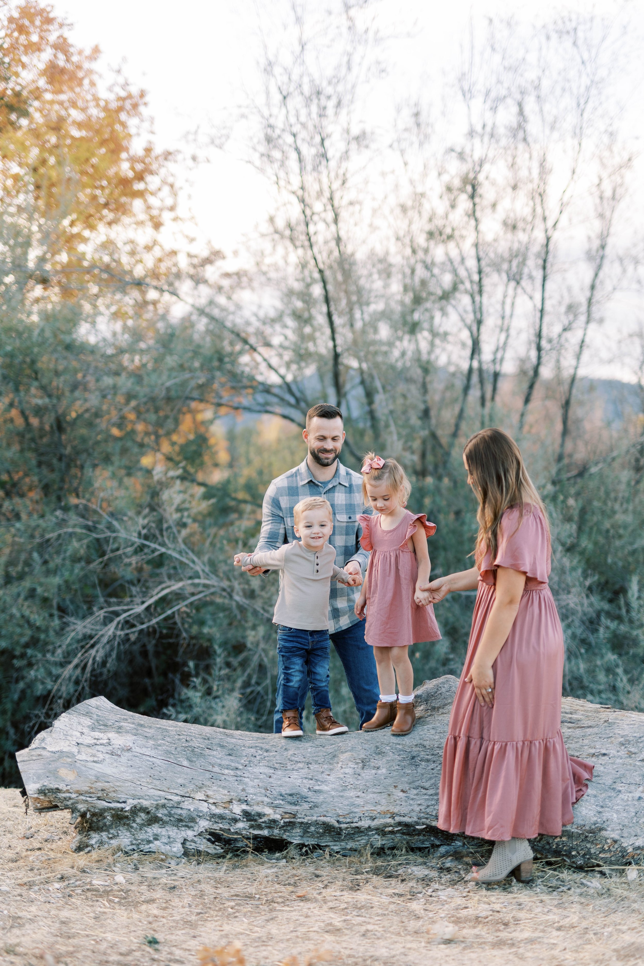 A family of four standing on a fallen tree in an outdoor setting during fall, with trees and sky in the background. The father is holding hands with a young boy and a young girl, while a woman, possibly the mother, stands nearby holding the young gir