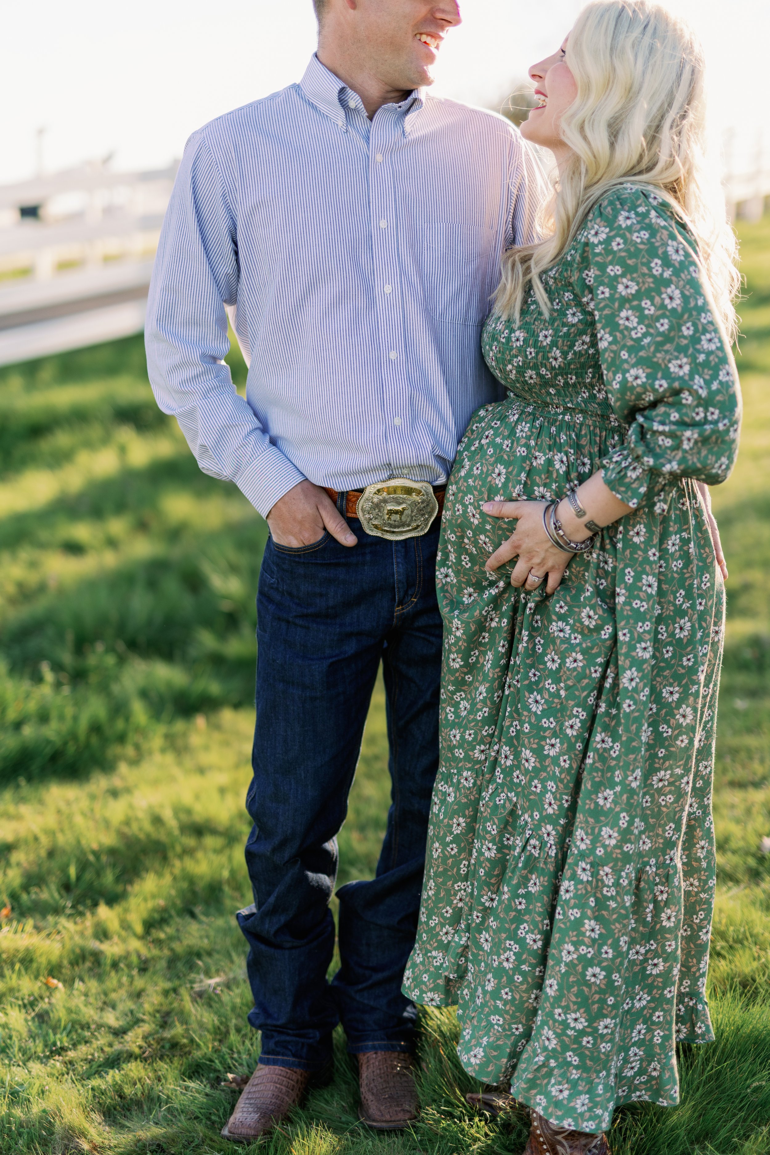 A pregnant woman standing next to a man outdoors, both smiling and looking at each other, with the woman touching her baby bump. The man is wearing a striped blue button-down shirt, jeans, and cowboy boots, with a large belt buckle. The woman has lon