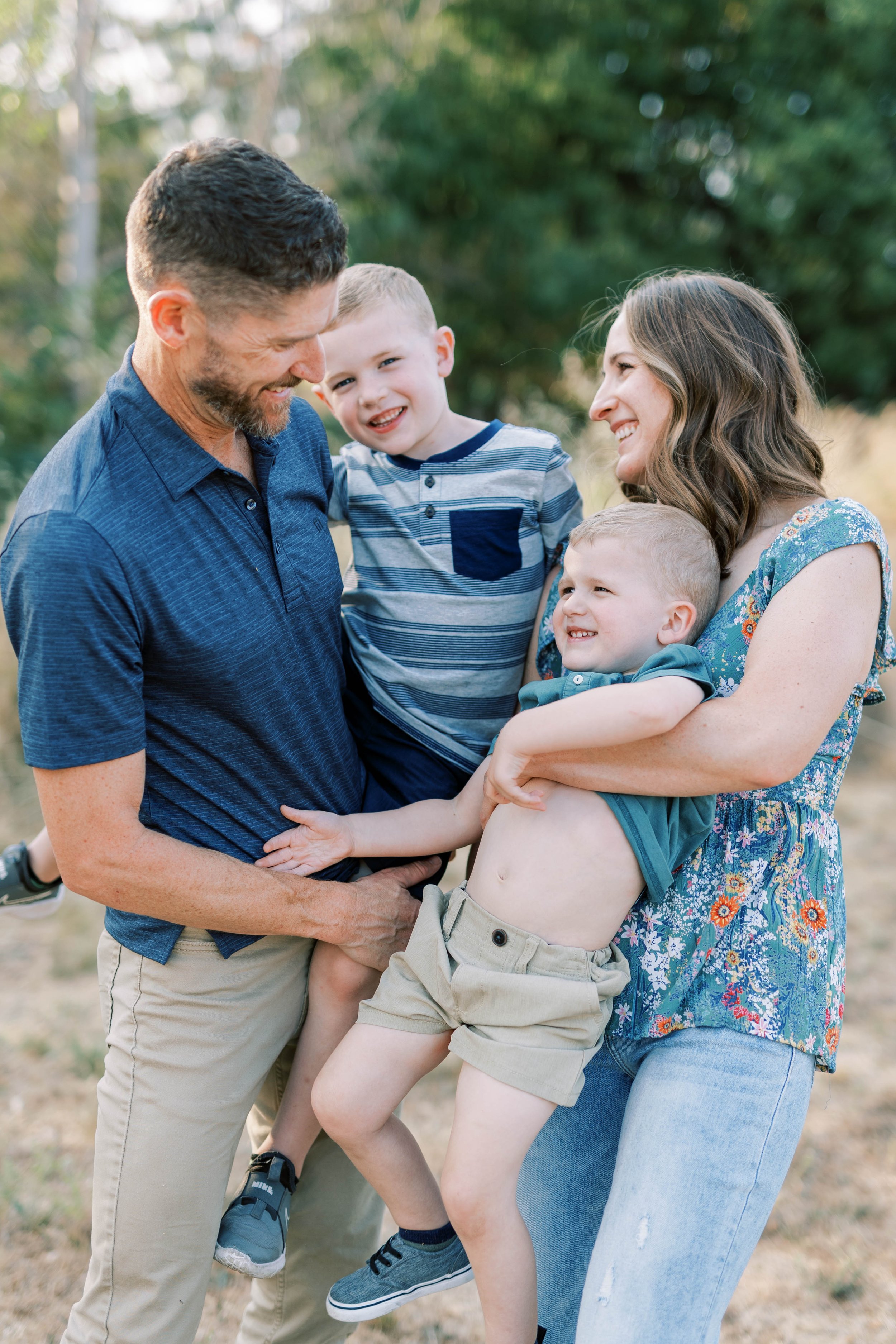 A happy family of four outdoors, with a father, mother, and two young boys, smiling and playing together. Family photography in Medford, Oregon.