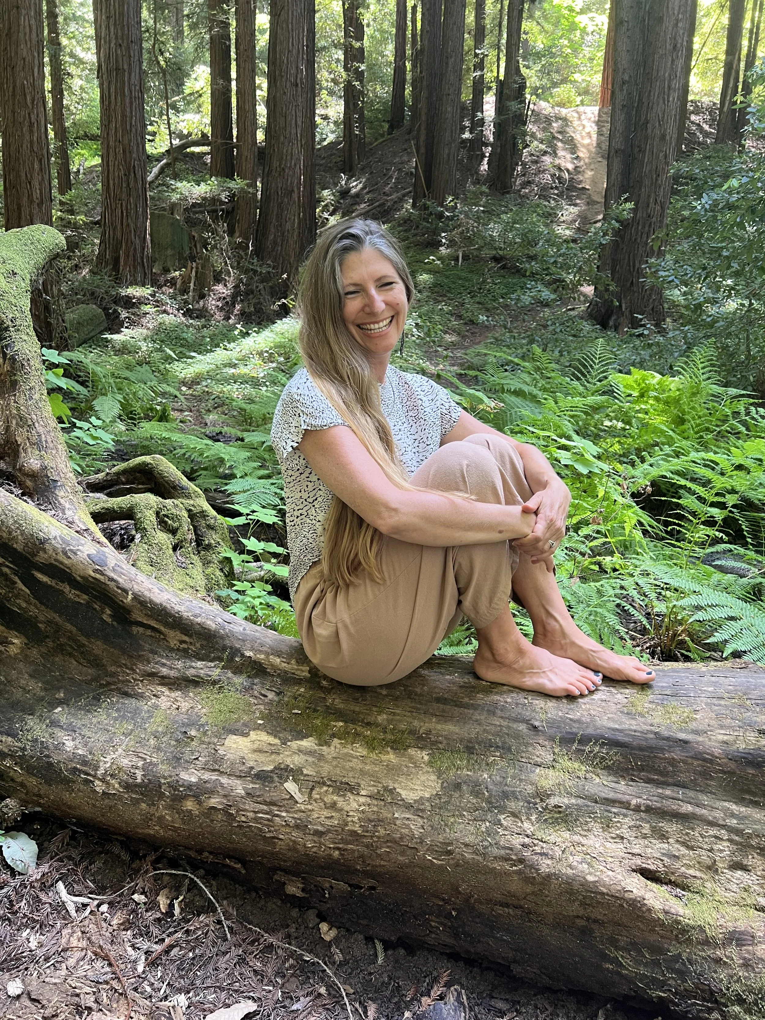 Amarisa with a huge smile sitting on a log hugging her knees lightly, bum and barefeet rooting her to the forest floor in pure delight.