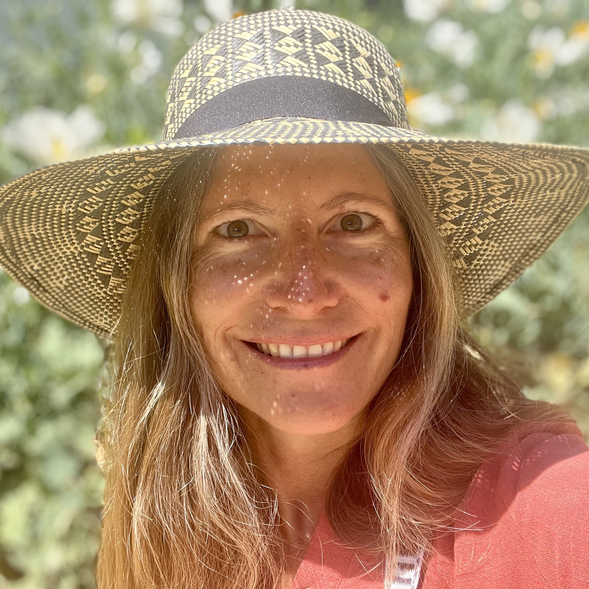 A portrait of Amarisa smiling in the sun with a wearing an intricately woven womu straw hat hand-woven by the Indigenous Wayuu peoples in front of a blurred background of the large white flowers of the Matilija Poppy.
