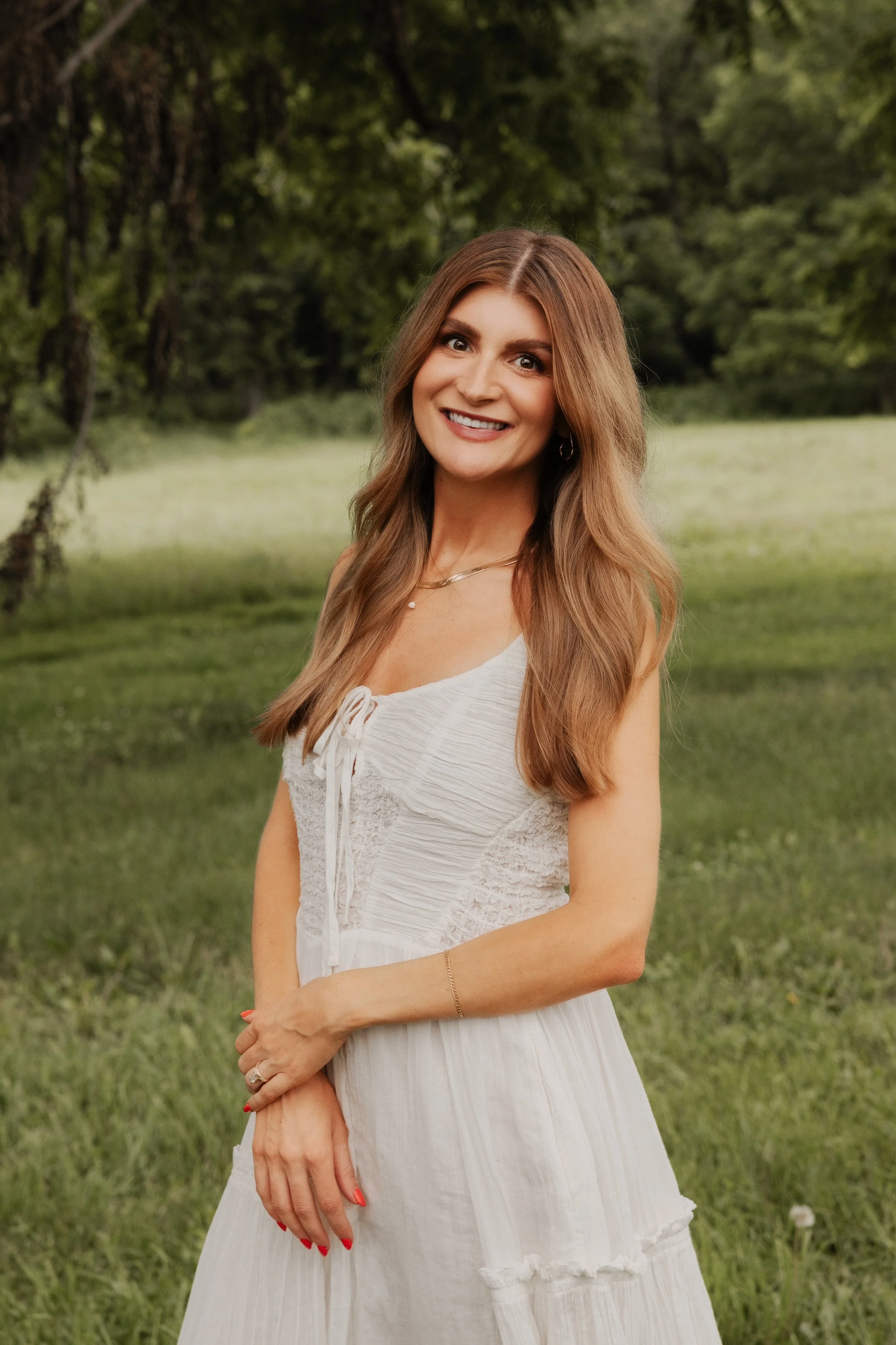 Woman with wavy brown hair wearing a beige sleeveless dress sitting on a wooden stool against a plain background.