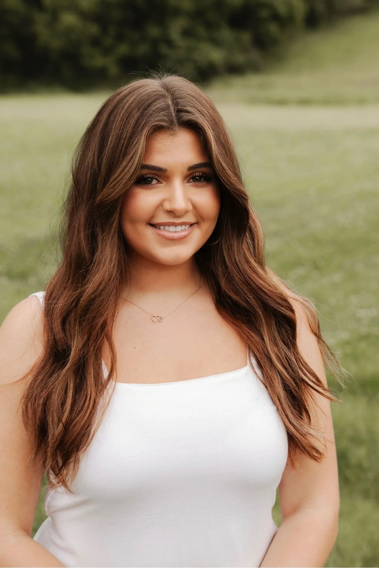 A woman with long dark hair, wearing a white denim jacket over a beige knit dress, smiling and posing against a neutral background.