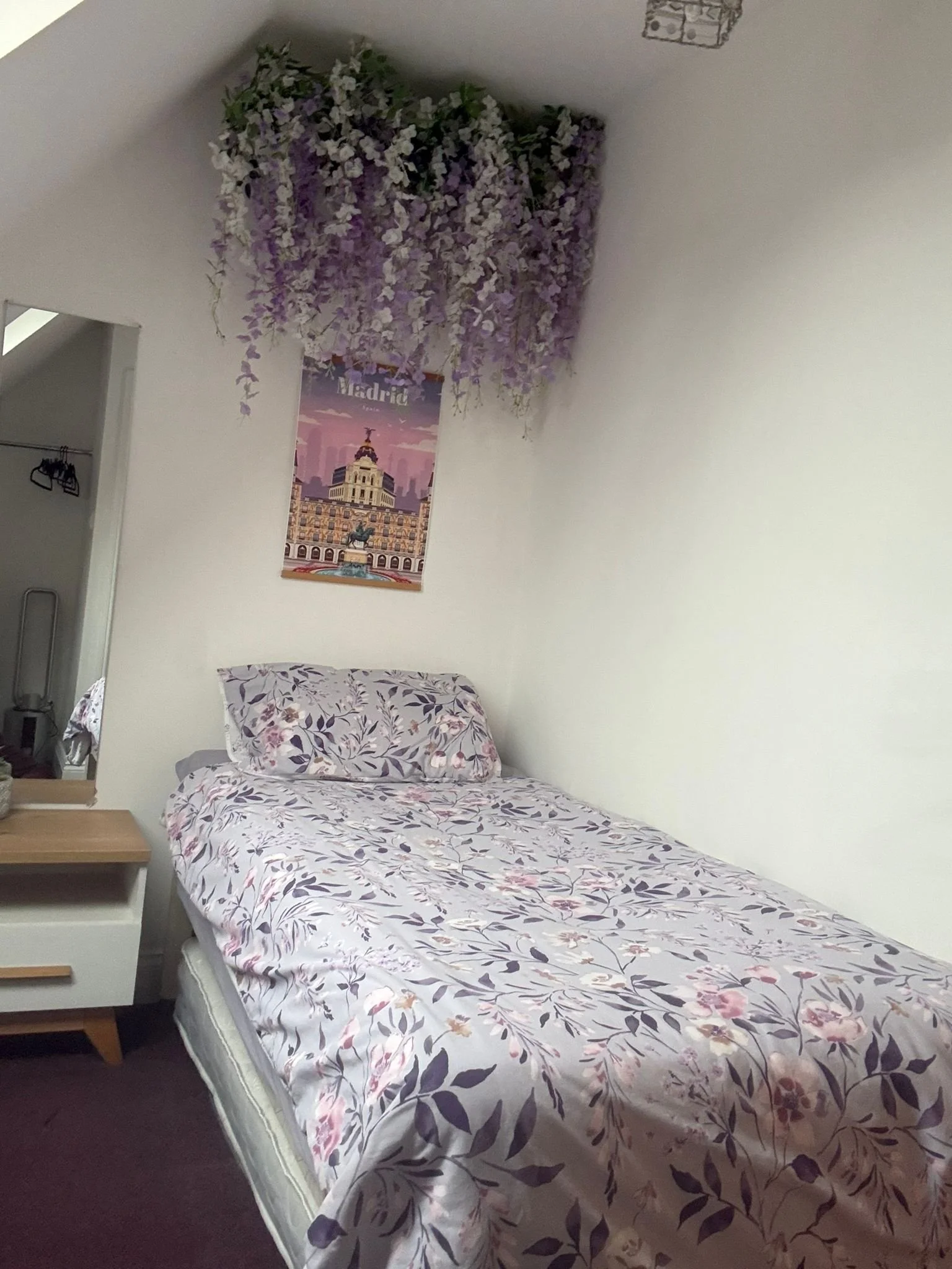 A bedroom with a bed covered in floral bedding, a matching pillow, a small wooden bedside table, and a white wall decorated with a Madrid poster and a hanging arrangement of purple and white flowers.