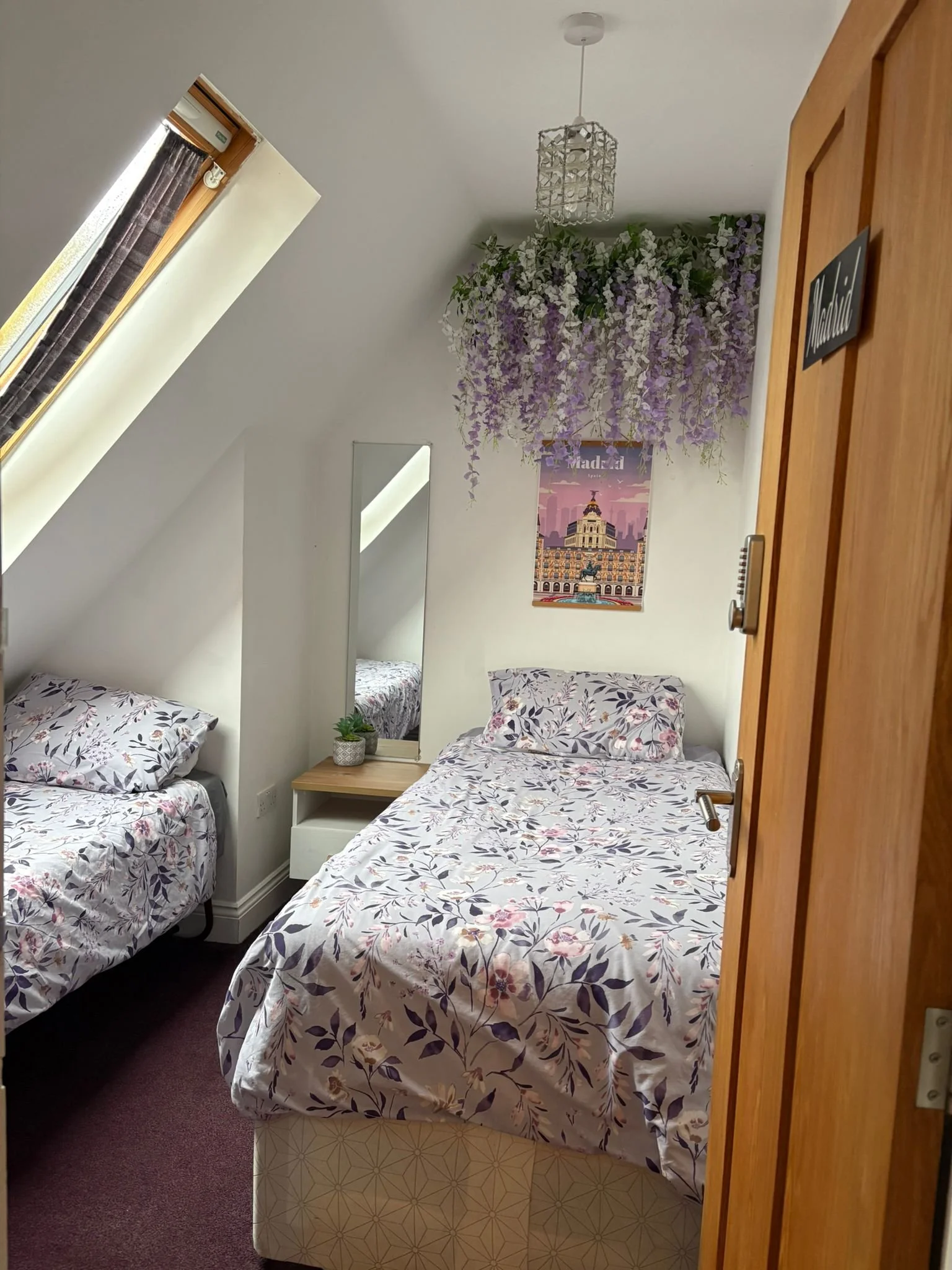 A small bedroom with two beds, one on the left and one on the right, with floral bedding. A narrow mirror and a small side table with a potted plant are between the beds. There's a window with a skylight on the sloped ceiling, purple flowers hanging 