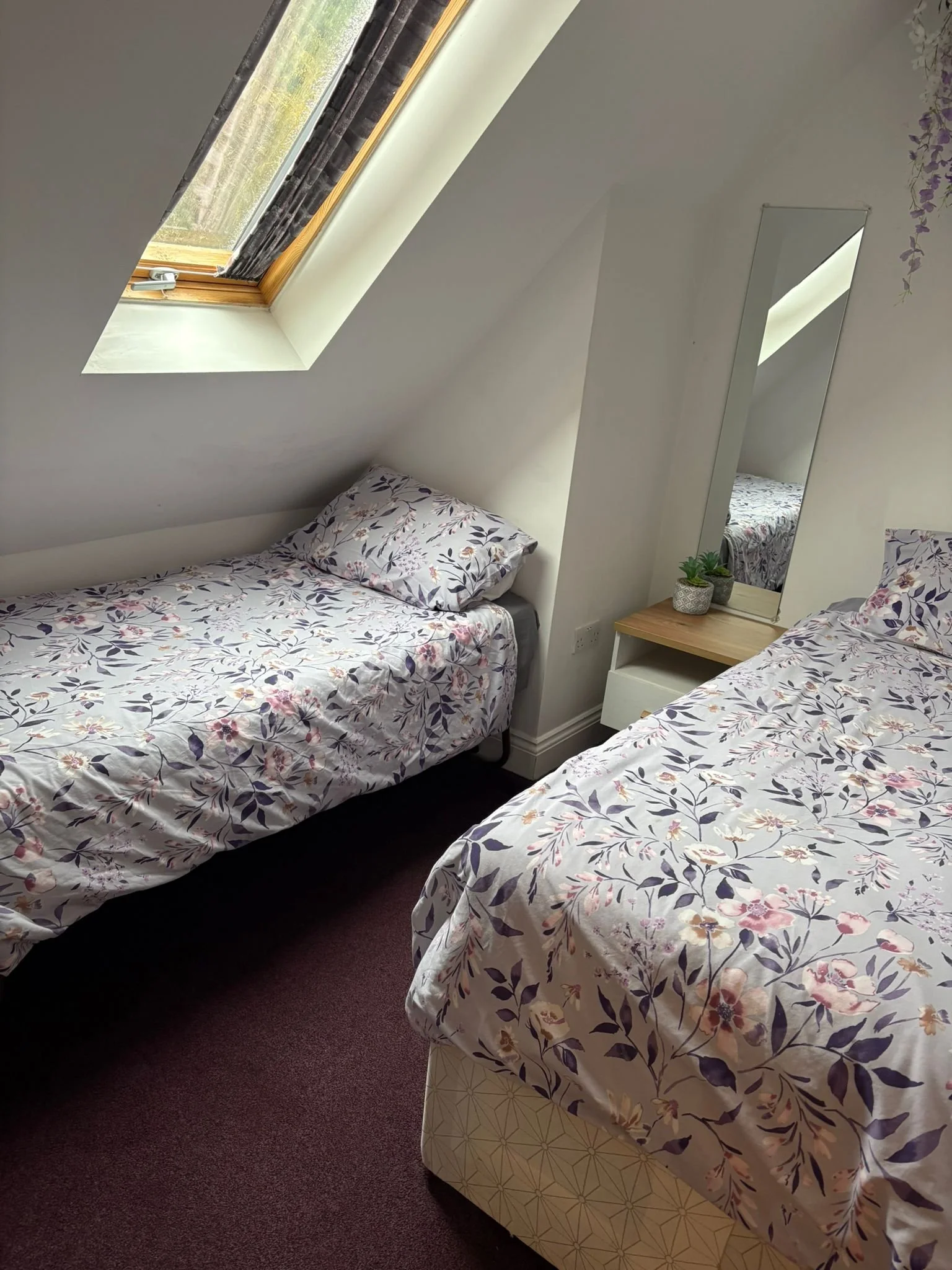 A bedroom with two single beds with floral bedspreads, a small nightstand with plants, a tall mirror, and a skylight window on a sloped ceiling.